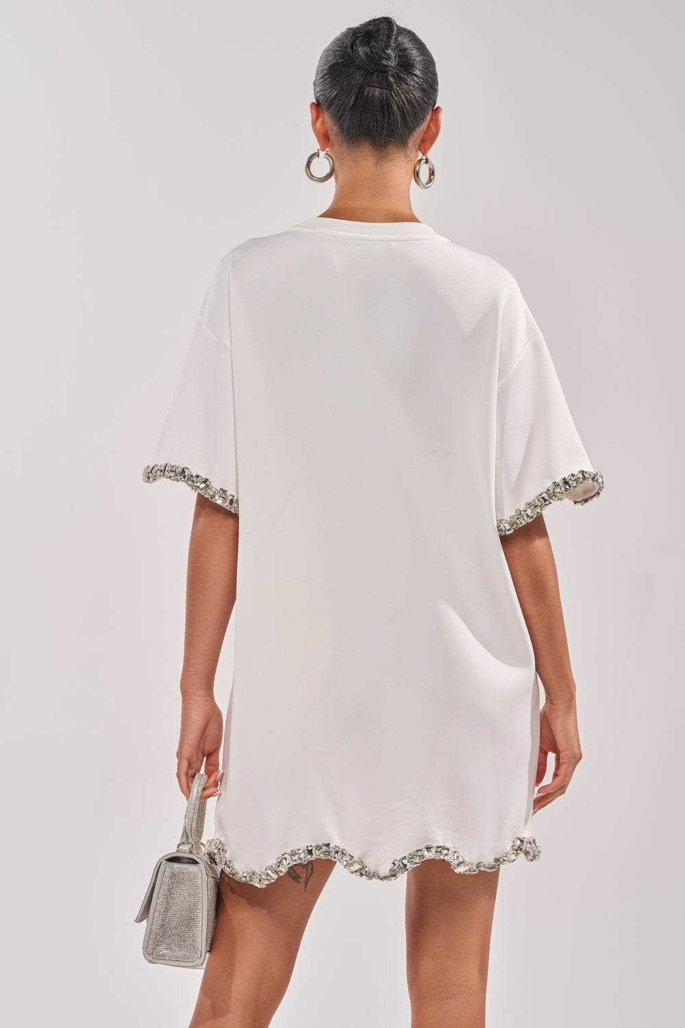 A person is seen from the back wearing the COOL GIRL EMBELLISHED TEE SHIRT DRESS IN WHITE—a loose mini dress with short sleeves, silver sequin trim, paired with a small silver handbag and large hoop earrings. 4