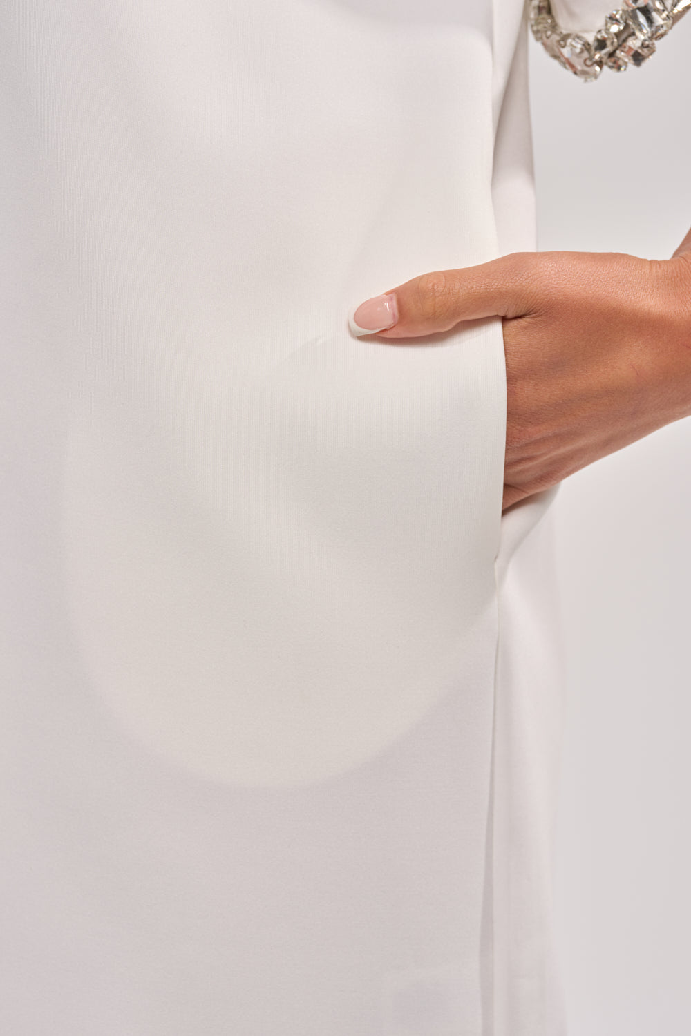 A close-up of a person’s right hand with manicured nails partially tucked into the pocket of the COOL GIRL EMBELLISHED TEE SHIRT DRESS IN WHITE, showcasing its soft, smooth fabric. 7