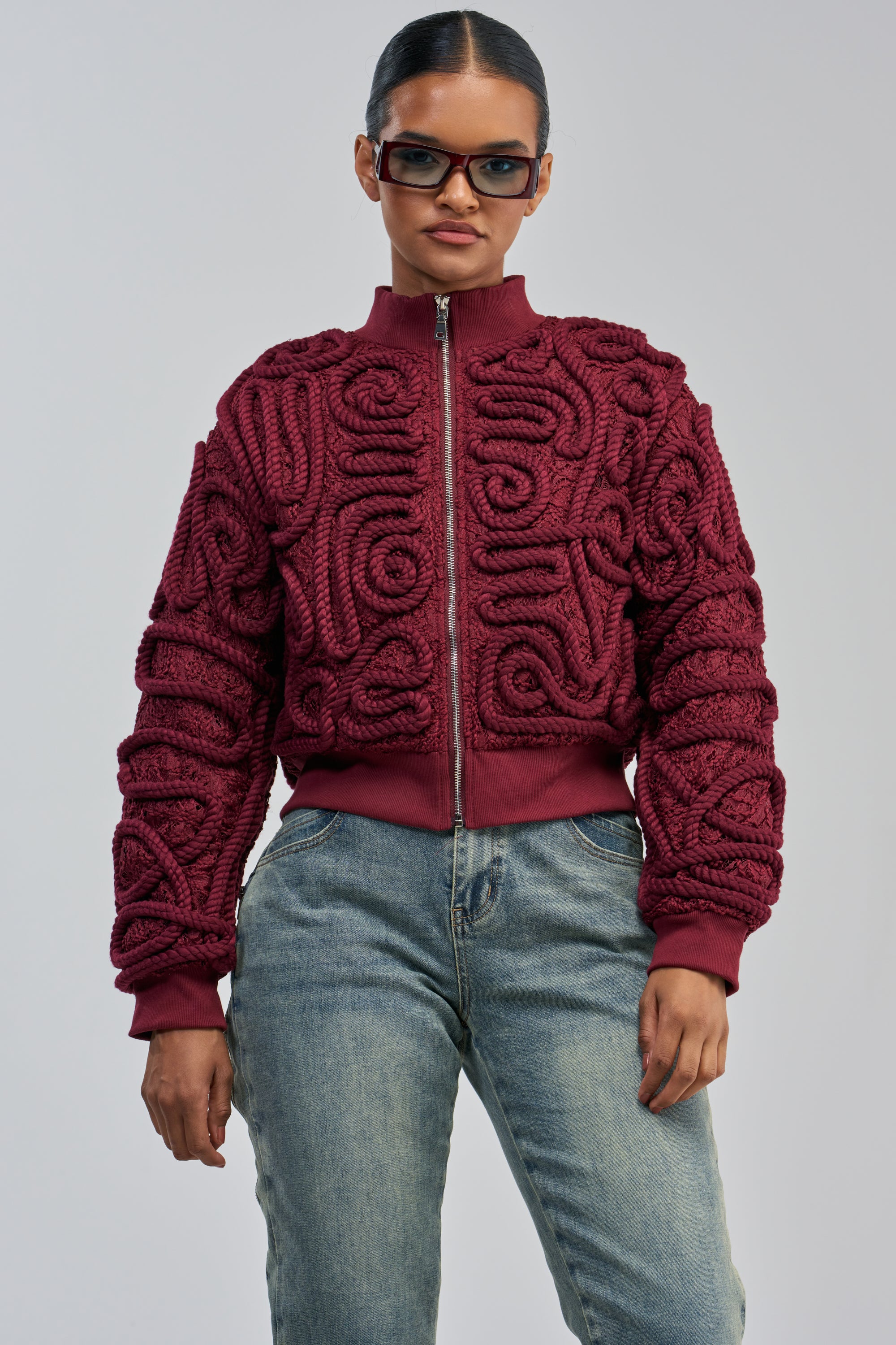 A person wearing sunglasses, light blue jeans, and the LOVER GIRL BOMBER—a statement streetwear burgundy lace bomber jacket with rope detail and raised patterns—stands against a plain gray background.