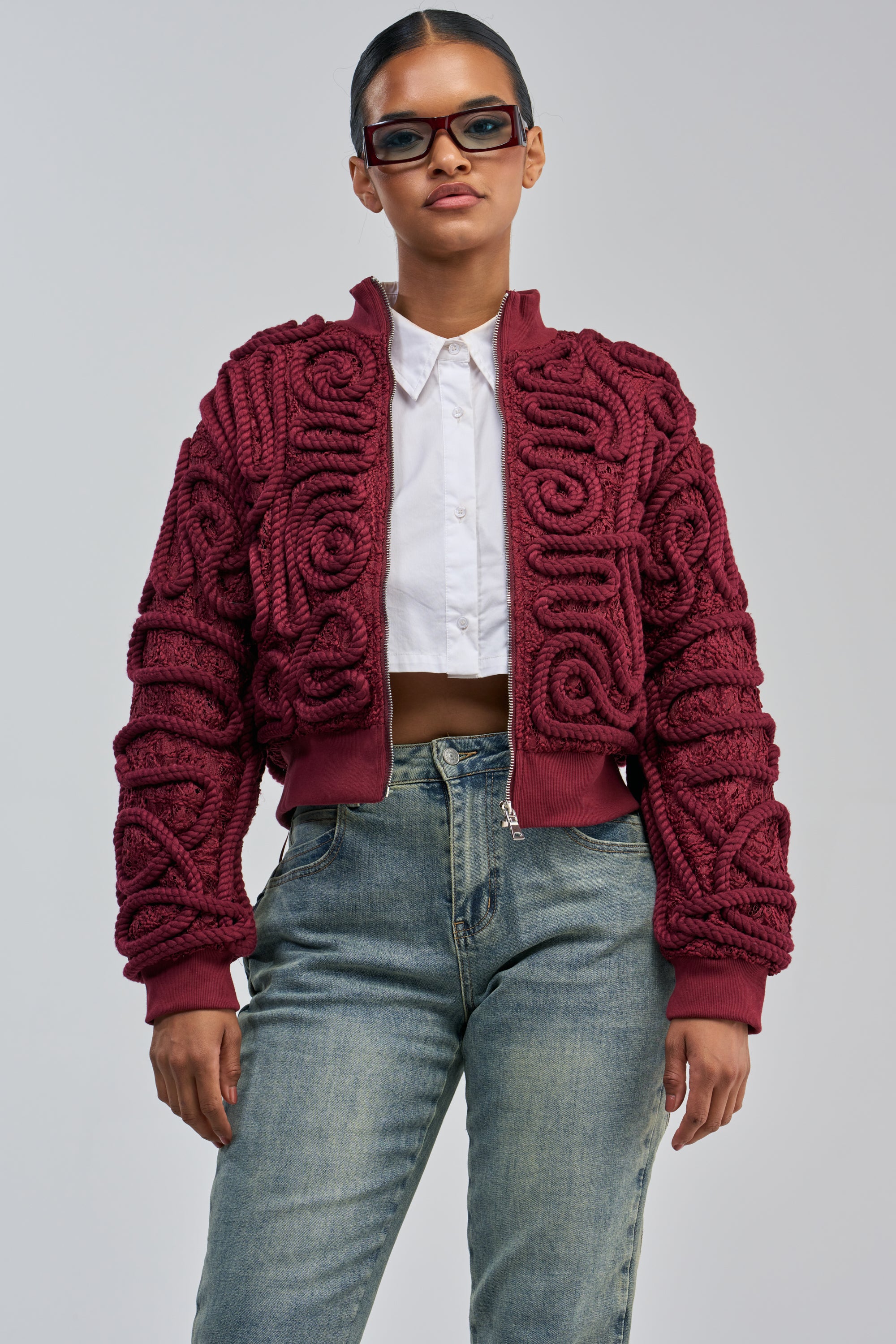 A woman in blue jeans, a white cropped shirt, and the LOVER GIRL BOMBER—a statement streetwear maroon jacket with a rope detail pattern—wears rectangular glasses while standing against a plain light gray background.