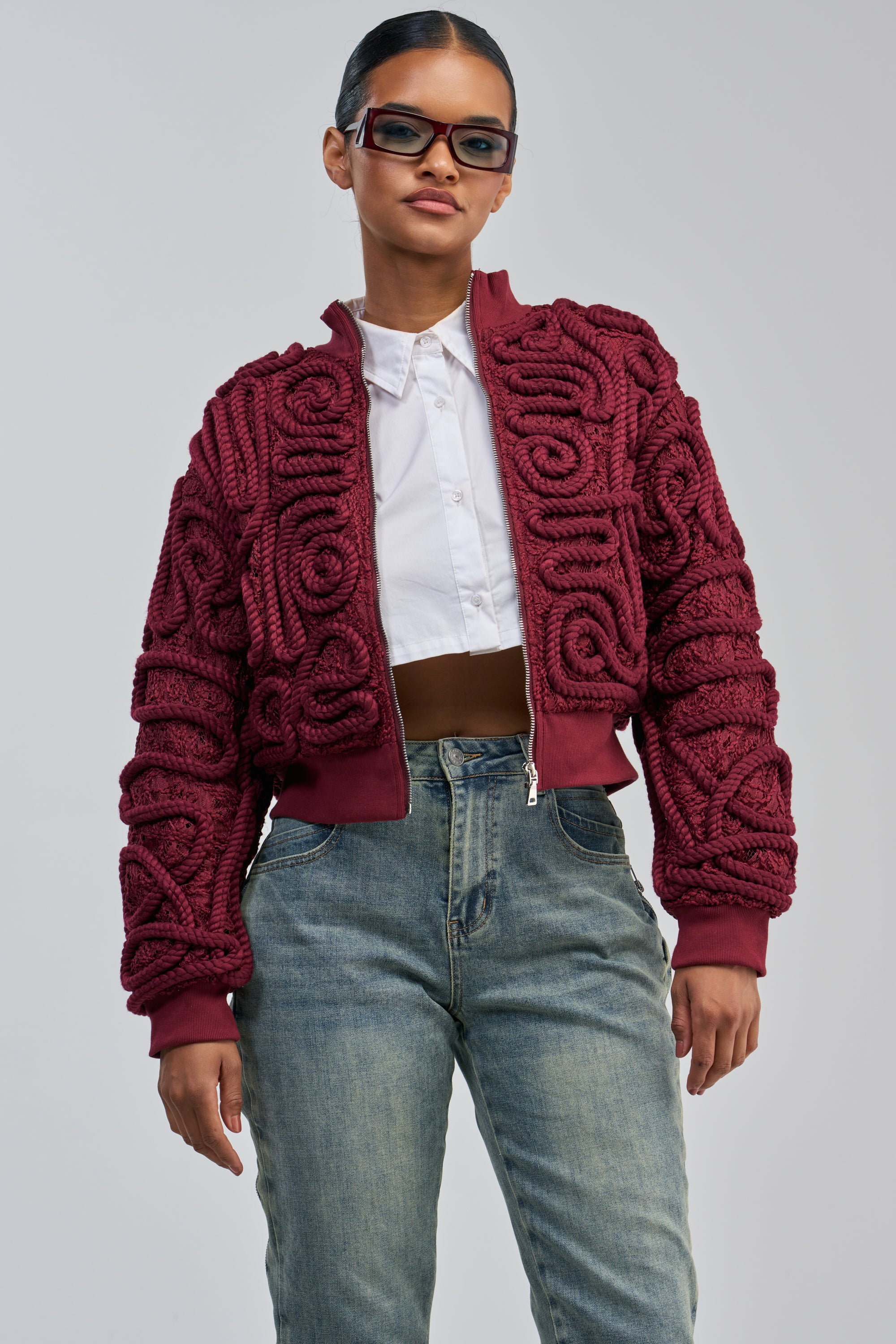 A woman stands confidently against a light gray background, embodying statement streetwear style in the LOVER GIRL BOMBER—a burgundy lace jacket with rope detail—paired with a white cropped shirt, blue jeans, and rectangular glasses.