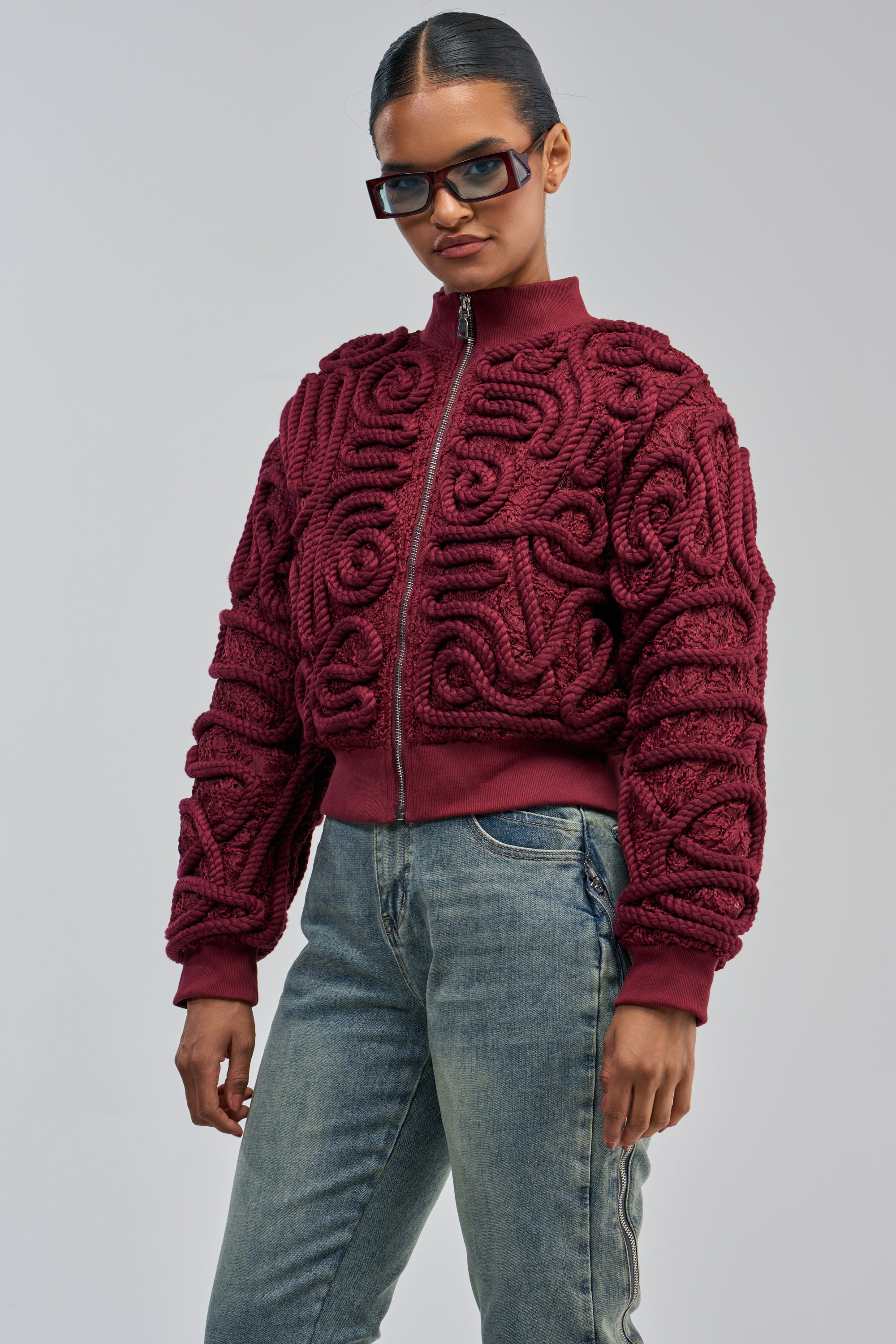 A person wearing dark glasses showcases a statement streetwear look featuring the LOVER GIRL BOMBER, a burgundy lace bomber jacket with intricate patterns and rope detail, paired with blue jeans, standing against a plain light gray background.