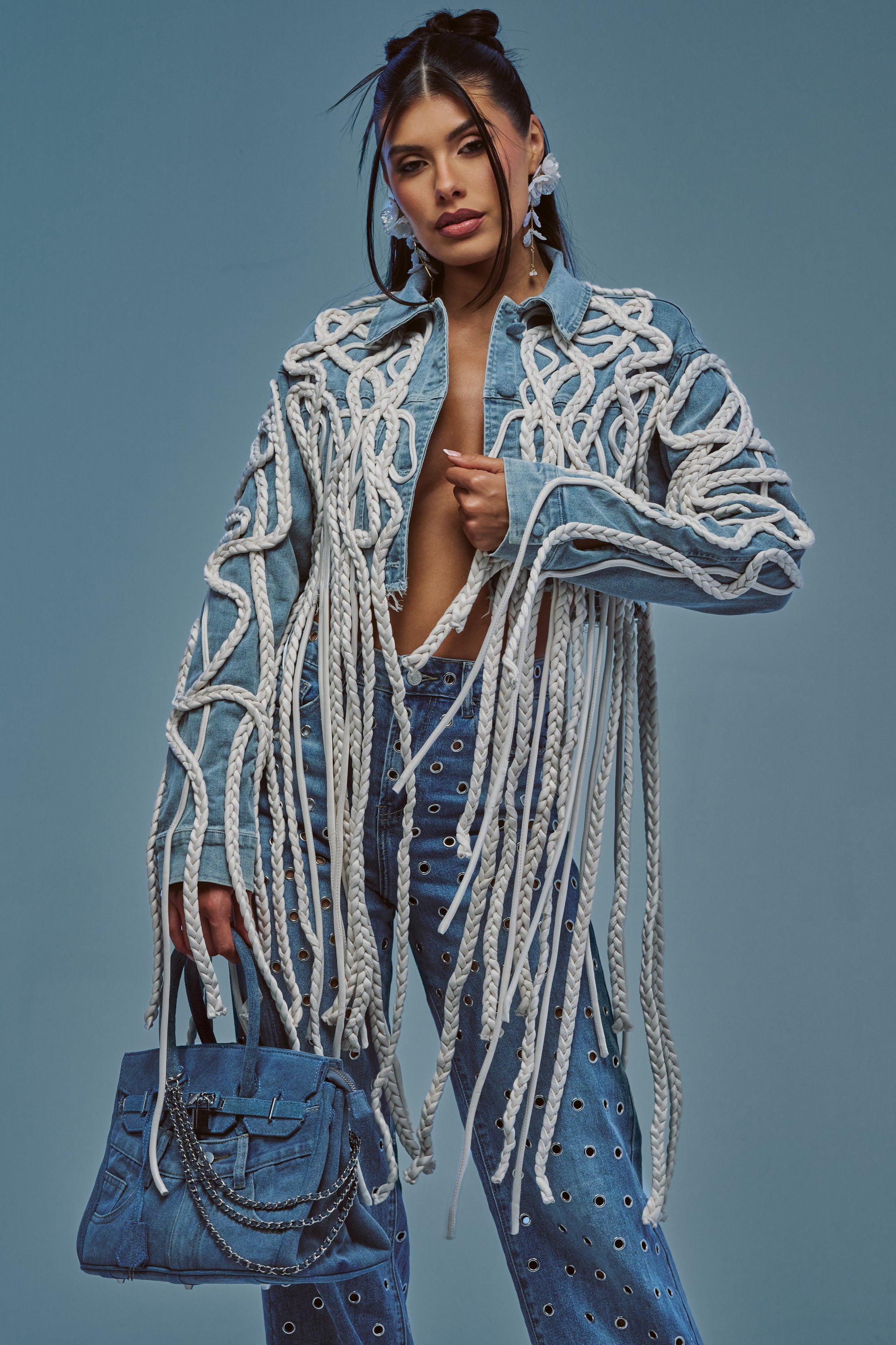 A woman poses against a plain blue background, wearing the KEEP MOVING TASSEL DENIM JACKET—an avant-garde streetwear piece embellished with thick white ropes and eyelet details—while holding a matching denim handbag with chain accents.