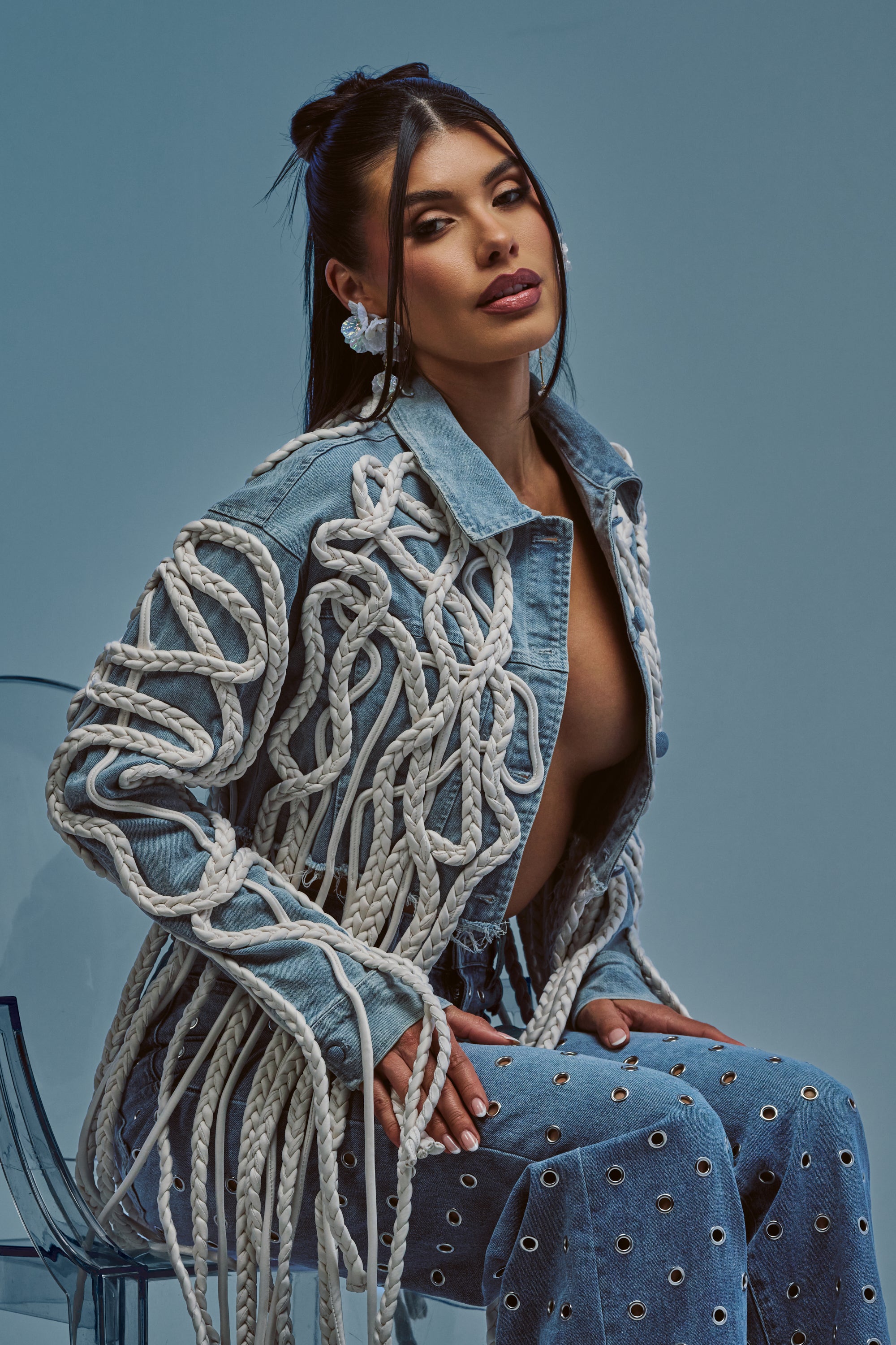 A woman sits on a clear chair, wearing the KEEP MOVING TASSEL DENIM JACKET—a unique streetwear-inspired denim piece with white rope details and grommet accents—against a blue background.