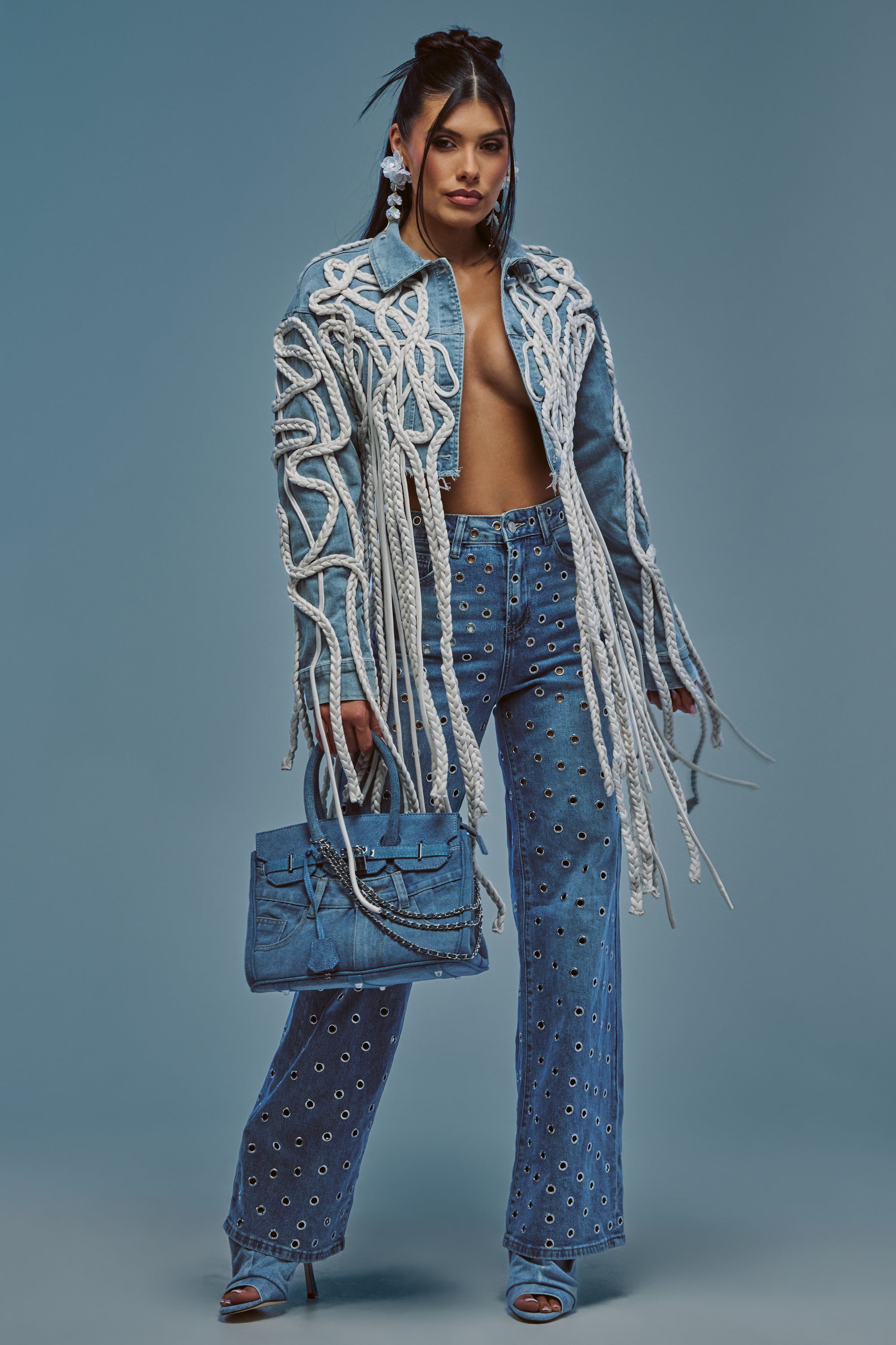 Wearing the KEEP MOVING TASSEL DENIM JACKET layered over a long denim coat with white rope details, embellished denim pants, blue heels, and carrying a blue handbag, a woman stands confidently against a blue background, embodying modern streetwear style.