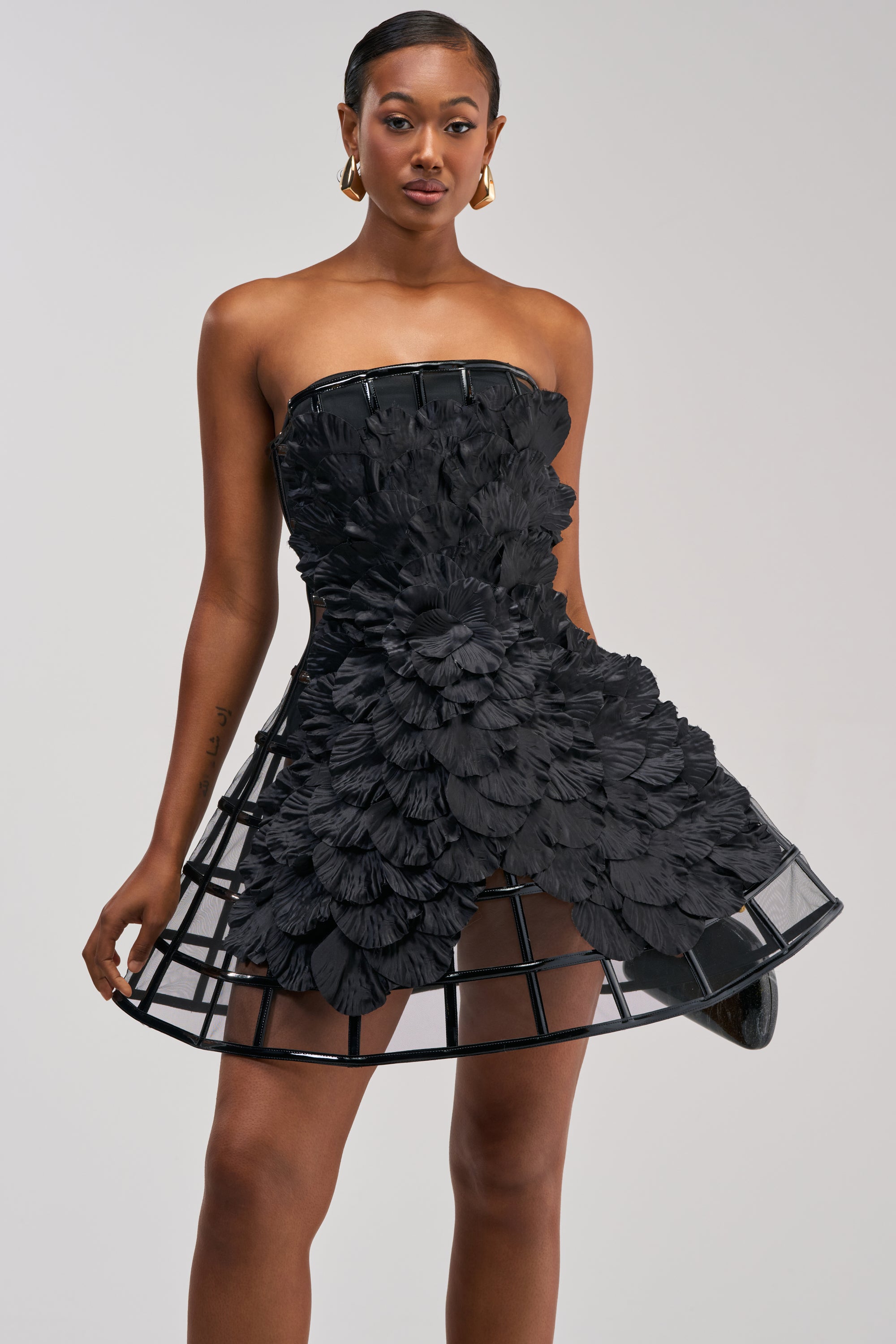 A woman wearing the FULL BLOOM CAGE DRESS IN BLACK—a strapless mini dress featuring 3D floral details and a structured, cage-like skirt—stands against a plain light background. This eye-catching piece is perfect as a statement cocktail party dress.