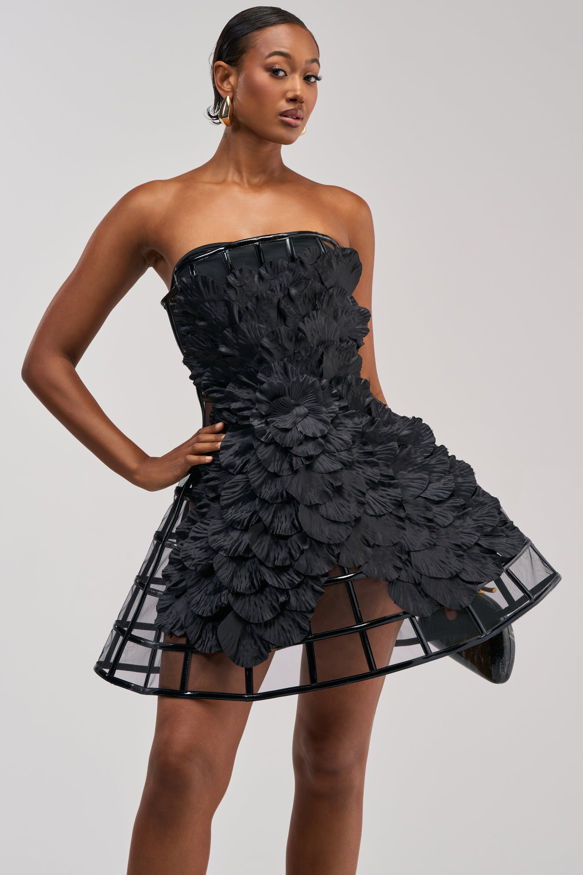 A woman models the FULL BLOOM CAGE DRESS IN BLACK, a chic mini dress featuring layered petal-inspired textures and a distinctive cage-like hem. Posing confidently with one hand on her hip against a plain backdrop, she effortlessly highlights this perfect cocktail party piece.