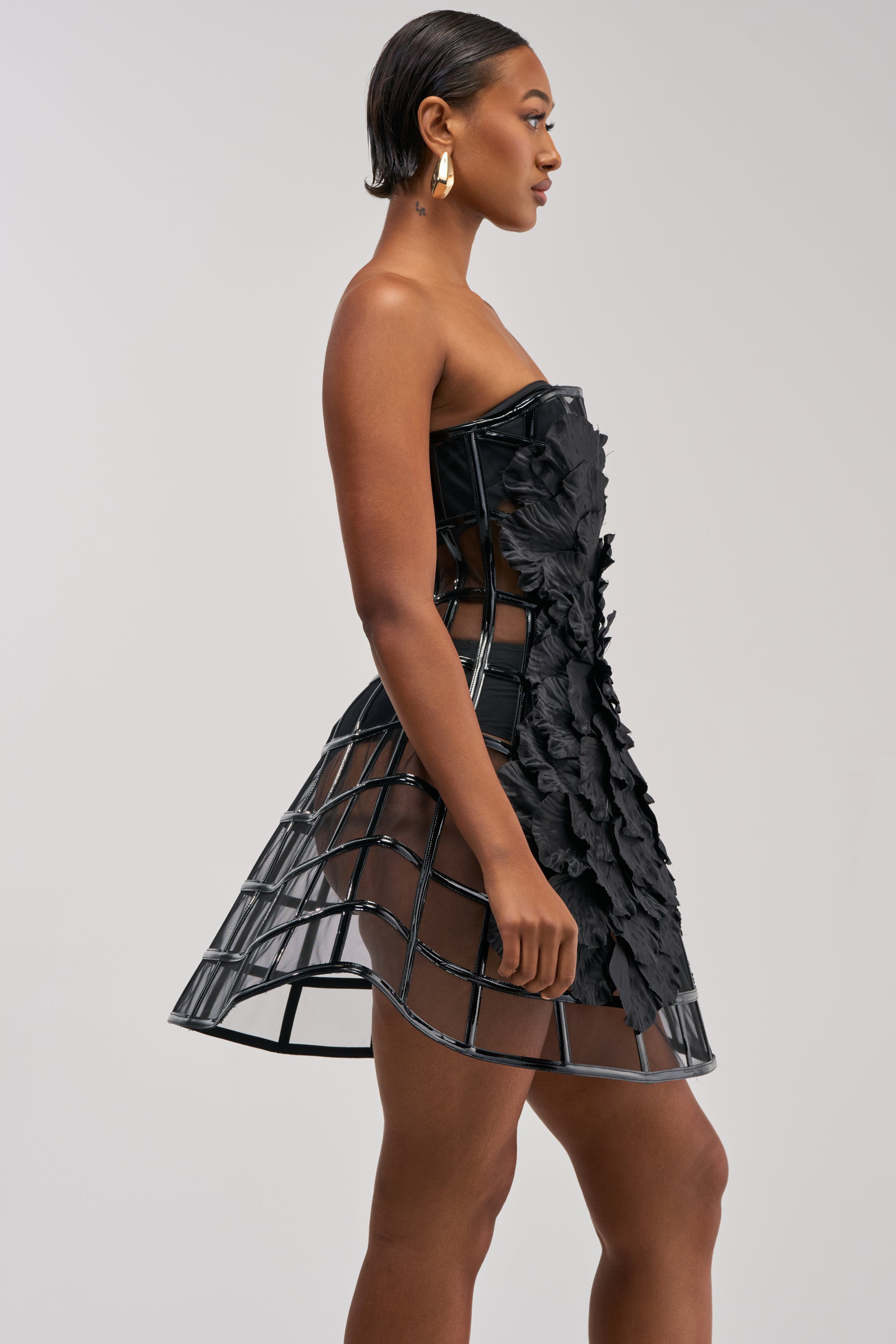 Wearing the FULL BLOOM CAGE DRESS IN BLACK, a woman poses sideways against a plain background, showcasing its structured cage-like skirt and delicate floral details—the perfect choice for a cocktail party.