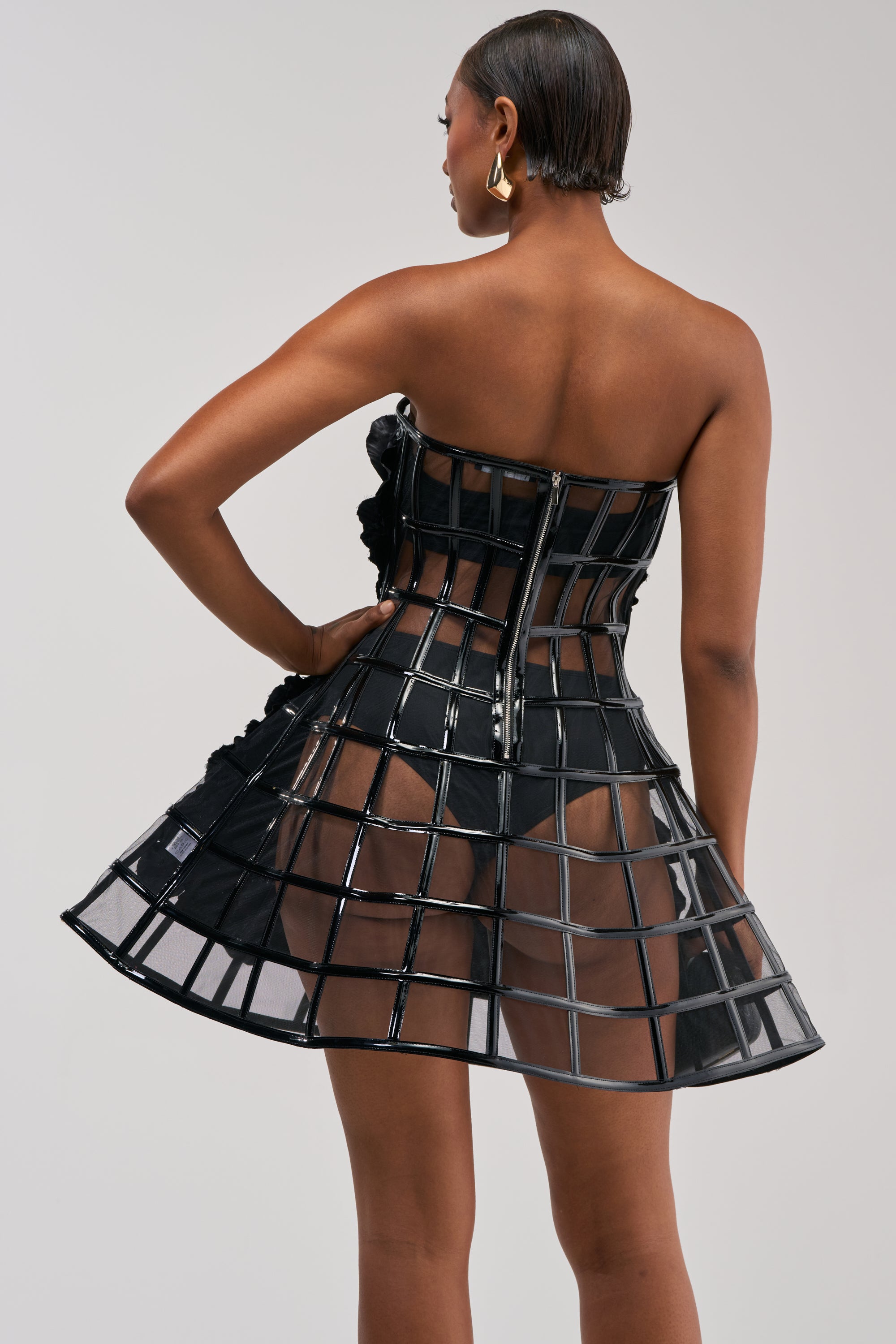 A woman stands with her back to the camera, showcasing the grid design and zipper closure of the FULL BLOOM CAGE DRESS IN BLACK, which features a strapless bodice and a structured, cage-like mini skirt.