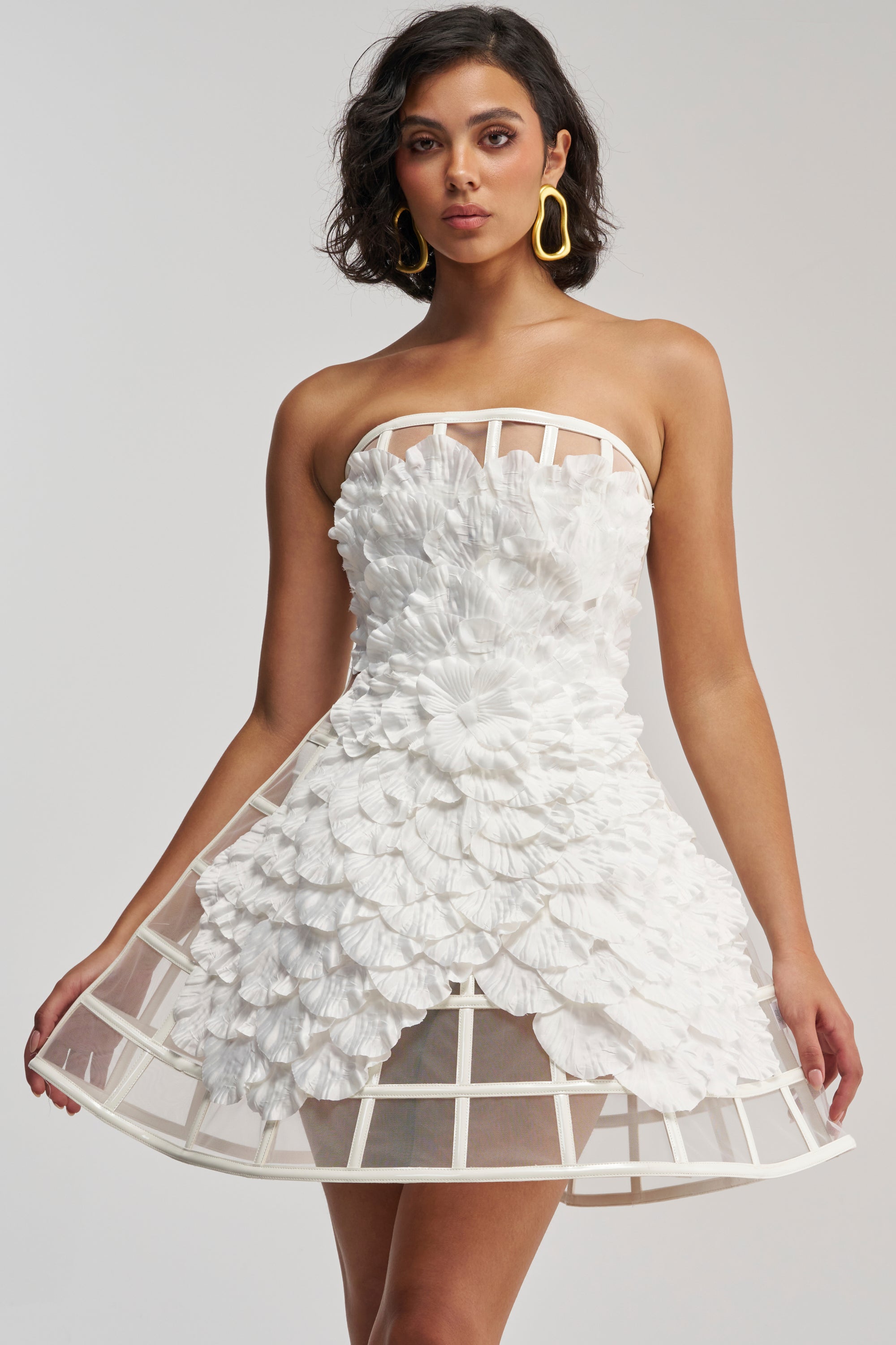 A woman poses against a plain light background, wearing the FULL BLOOM CAGE DRESS IN WHITE—a strapless mini dress featuring a white floral design with petal-like 3D textures and a distinctive cage structure at the hem. She completes her look with short dark hair and large yellow earrings.