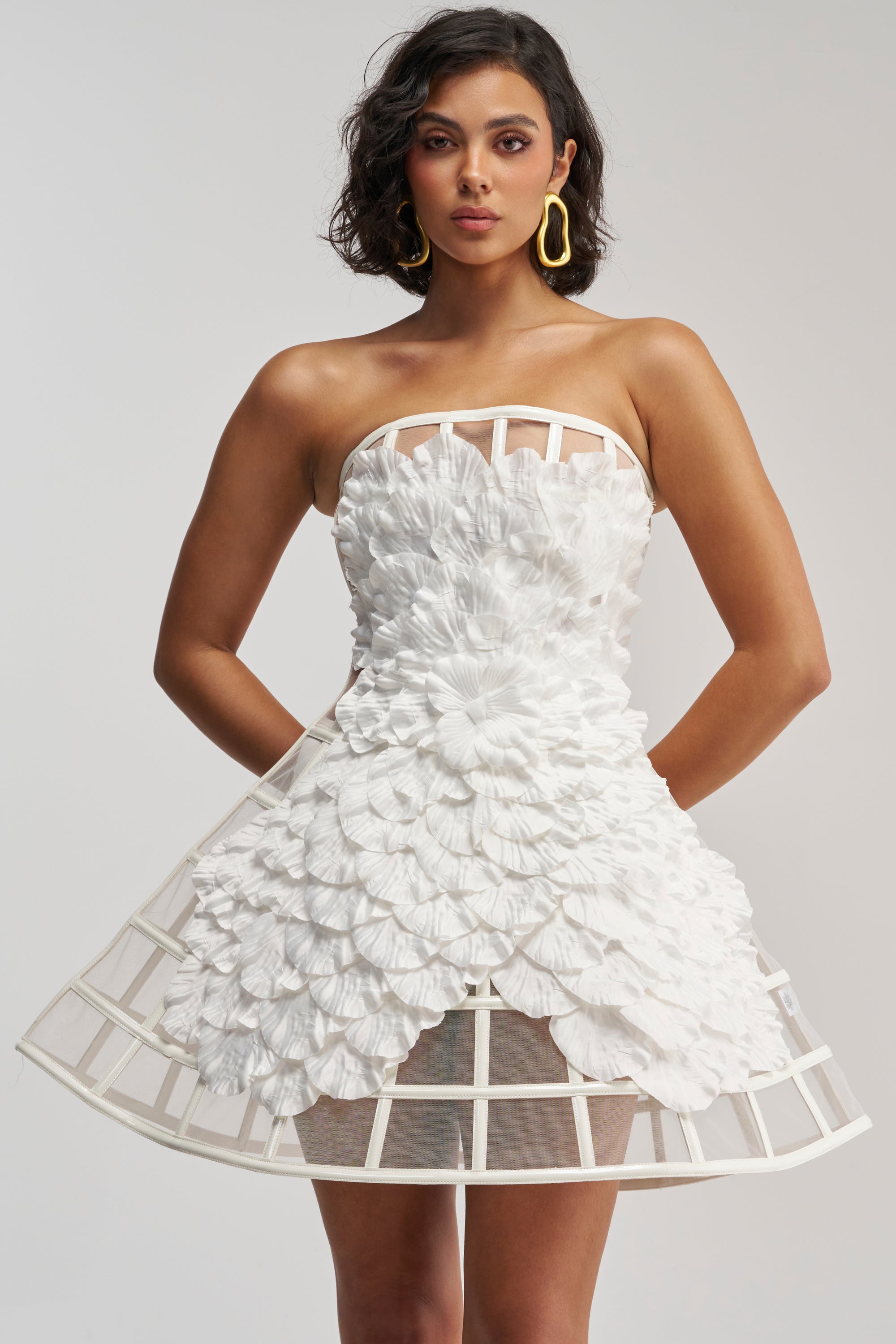 Model wears the FULL BLOOM CAGE DRESS IN WHITE, a mini dress featuring textured, petal-like details and a structured, cage-inspired skirt. She stands with hands in pockets and gazes confidently at the camera against a plain background.