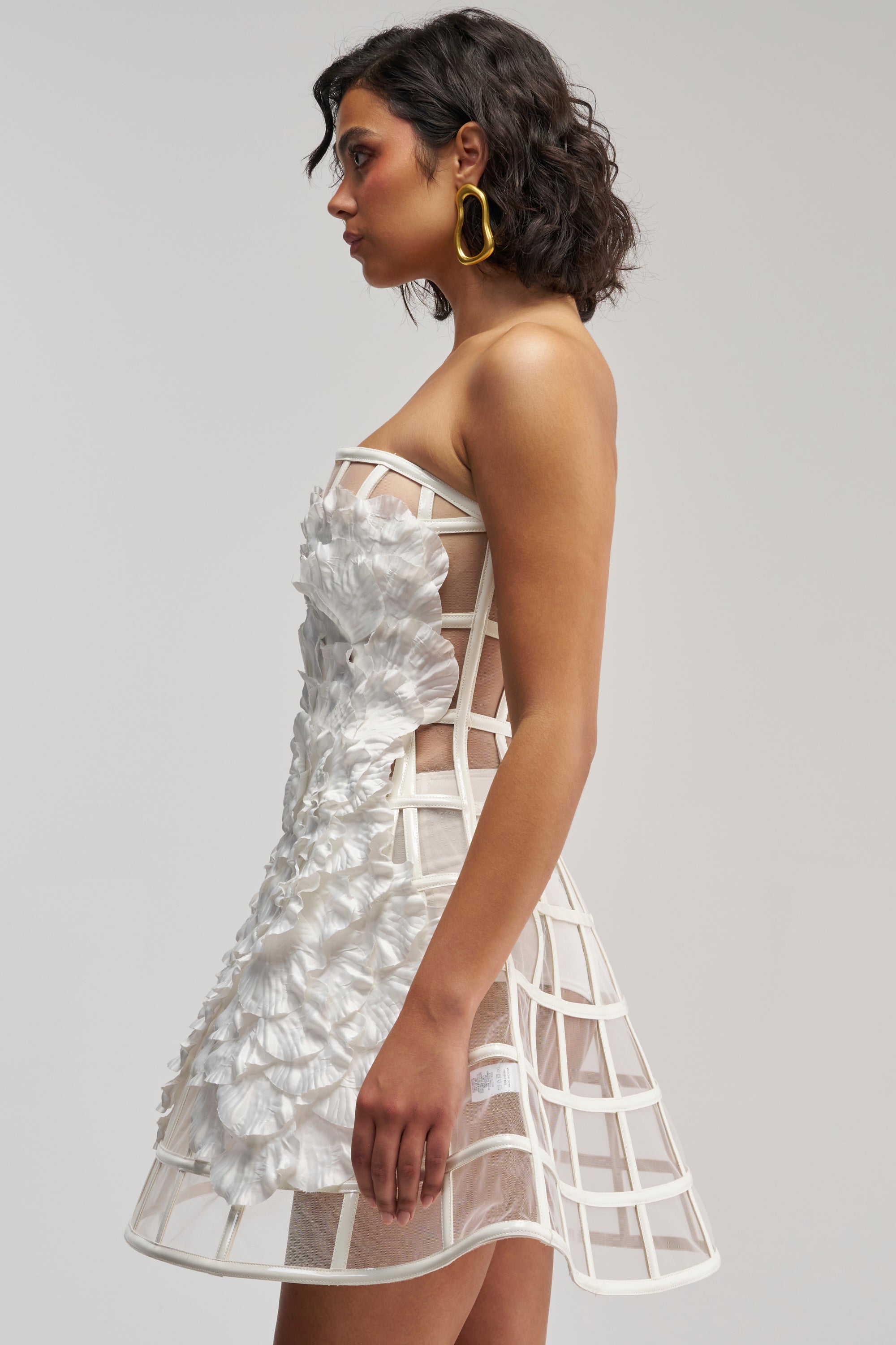 A woman in profile showcases the FULL BLOOM CAGE DRESS IN WHITE—a striking white mini dress featuring a cage-like grid structure adorned with delicate floral appliqués. Styled with large gold hoop earrings and set against a plain background, this unforgettable dress is the perfect modern choice for bridal parties.