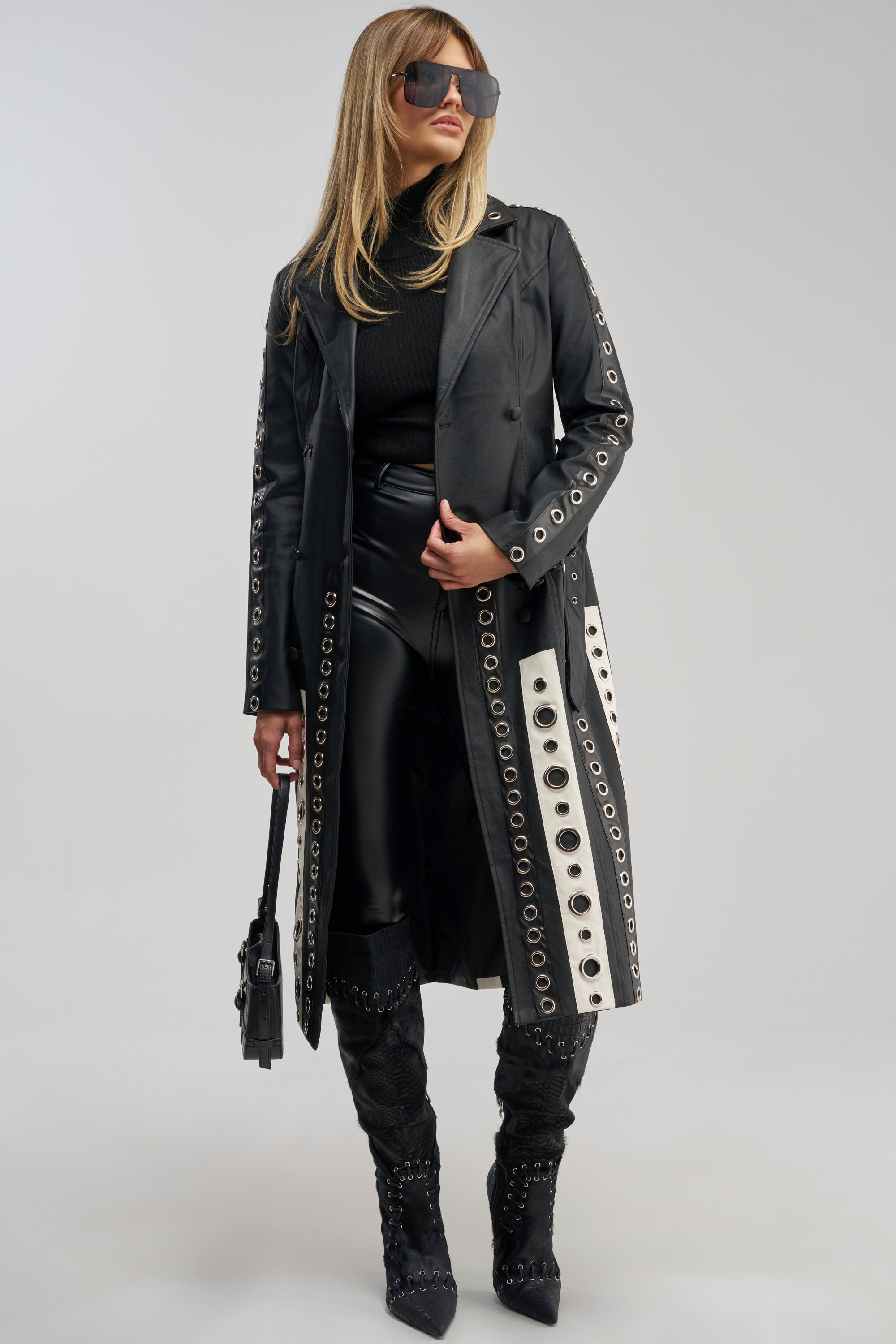 A woman wears the FIGURE IT OUT GROMMET TRENCH—a black faux leather trench detailed with silver grommets—styled with a black turtleneck, faux leather pants, black knee-high boots, sunglasses, and a black handbag, showcasing statement layering against a plain light background.