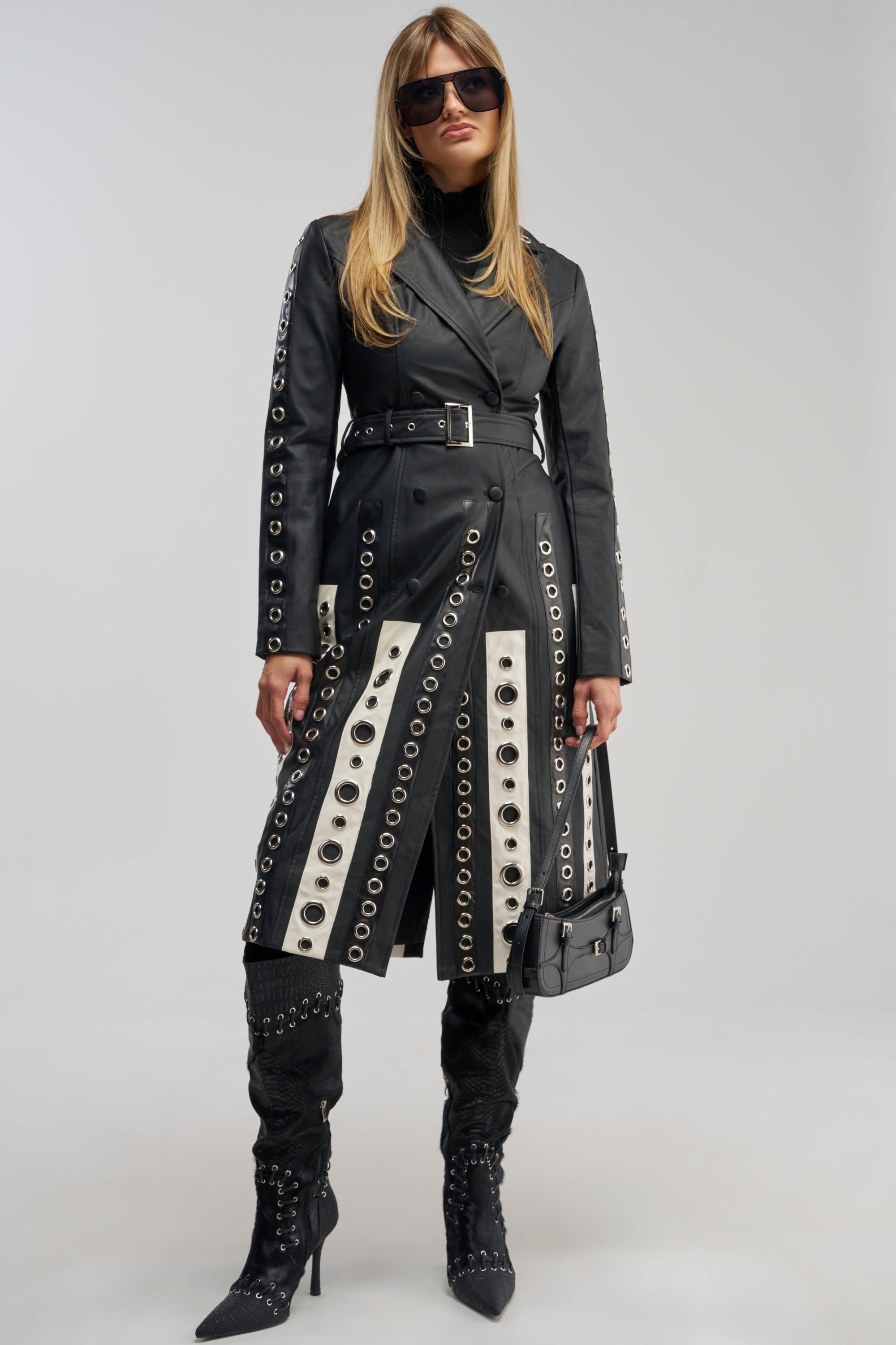 Wearing the FIGURE IT OUT GROMMET TRENCH, a woman stands against a plain background, showcasing statement layering with black boots and sunglasses. The black belted trench coat features silver grommet details, while she completes the look with a small purse.