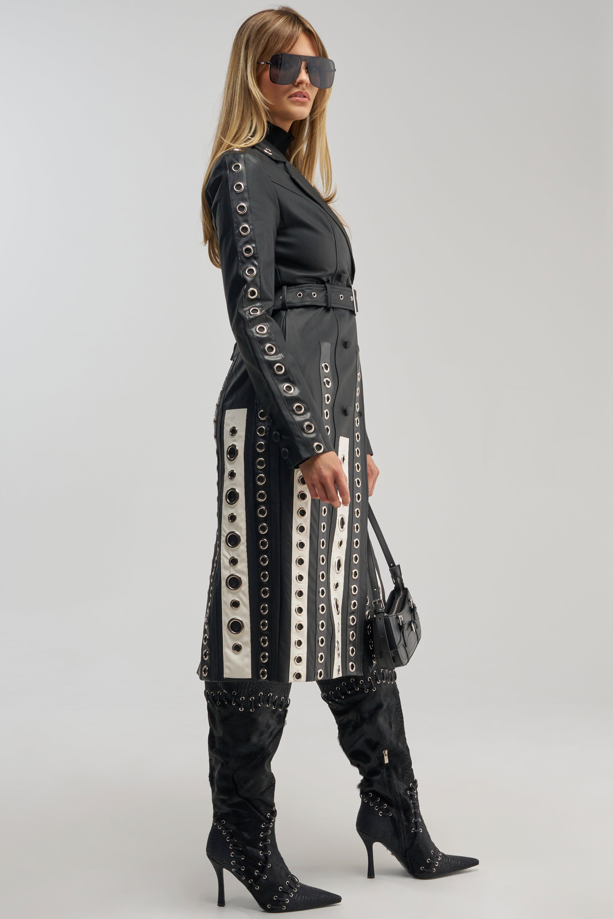 Wearing the FIGURE IT OUT GROMMET TRENCH—a black belted coat with bold grommet details—this woman pairs it with black high-heeled boots, oversized sunglasses, and a small purse as she stands against a light background.