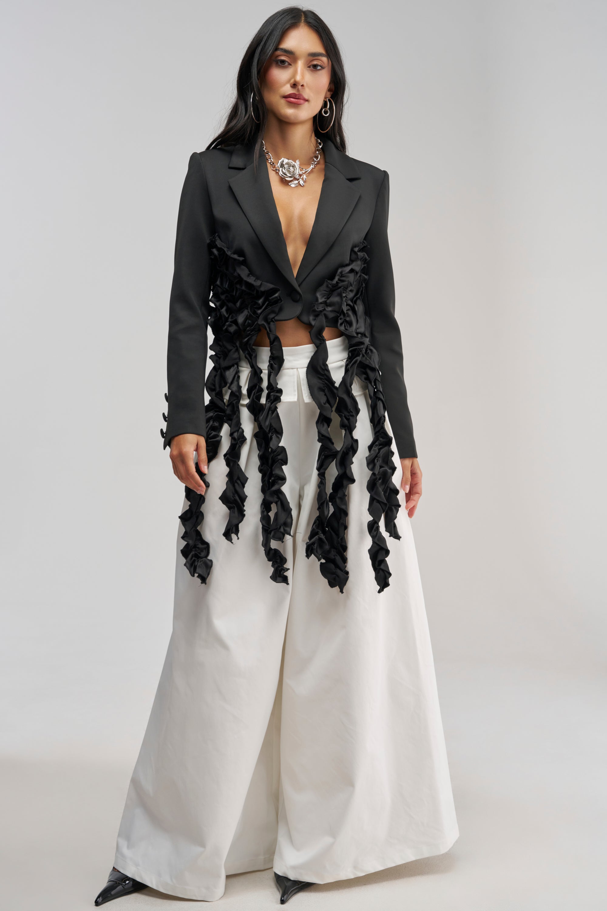 Wearing the CAUGHT YOUR EYE RUFFLE TRIM BLAZER paired with wide-leg white pants, statement jewelry, and black heels, a woman stands confidently against a plain background.