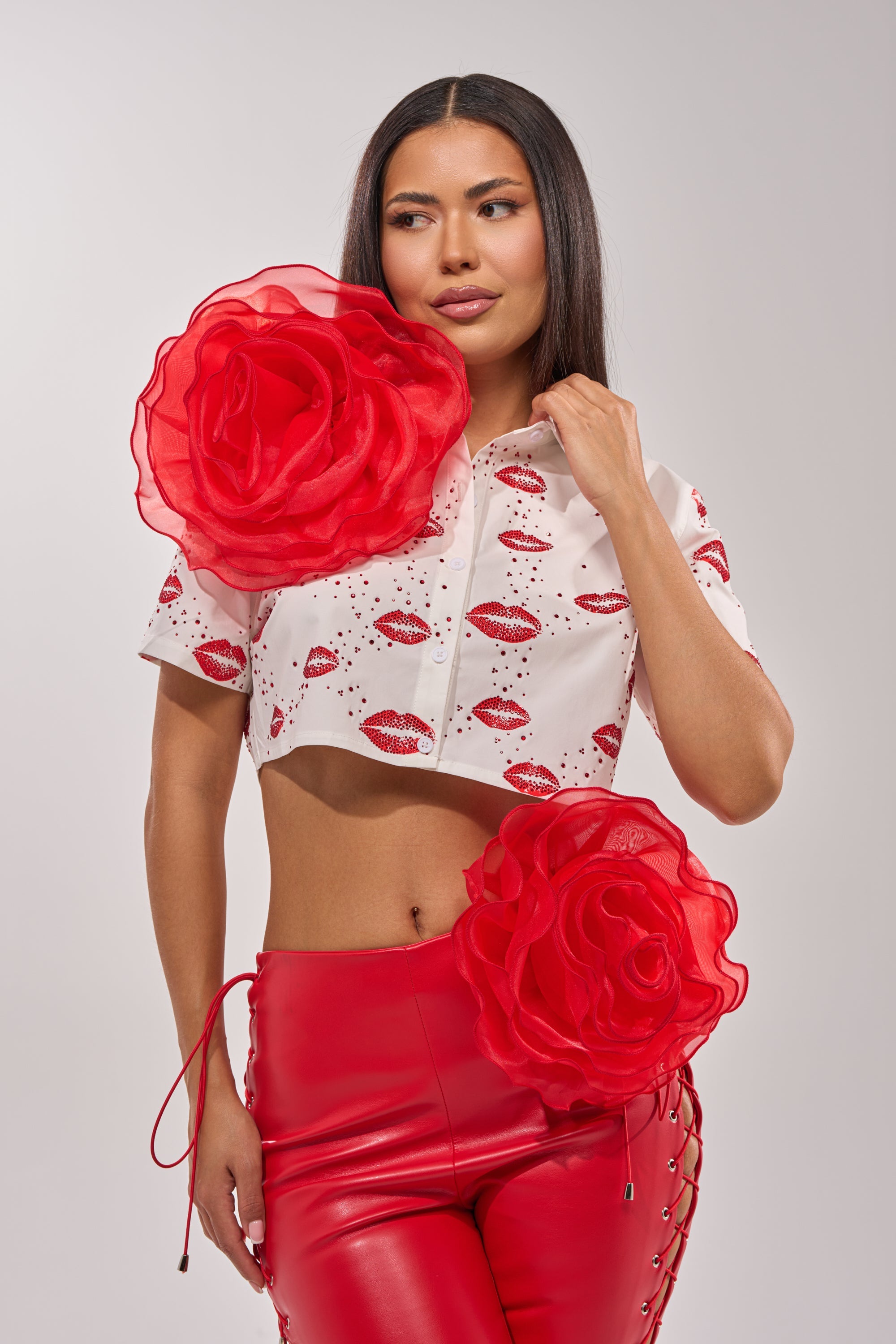 A woman wears the REDDY FOR YOU BROOCH SET: a white crop top with red lips print and an oversized floral brooch, styled with shiny red pants featuring a bold rose and lace-up sides, against a light background. 0