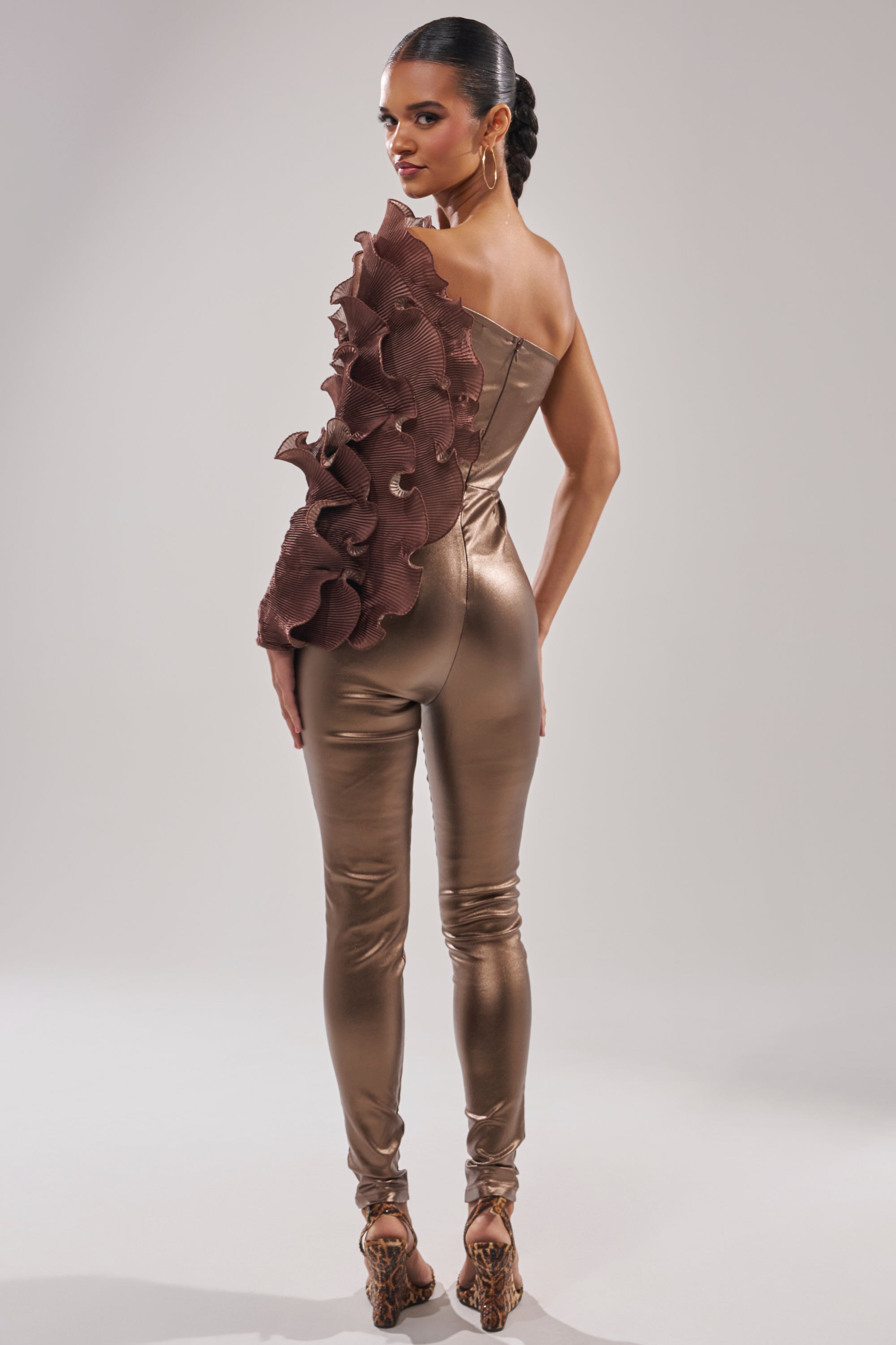A woman faces away, wearing the COCOA BUTTER ONE SLEEVE JUMPSUIT in metallic bronze with bold ruffle detail. Her hair is slicked back in a braided ponytail and she pairs the look with high heels.
