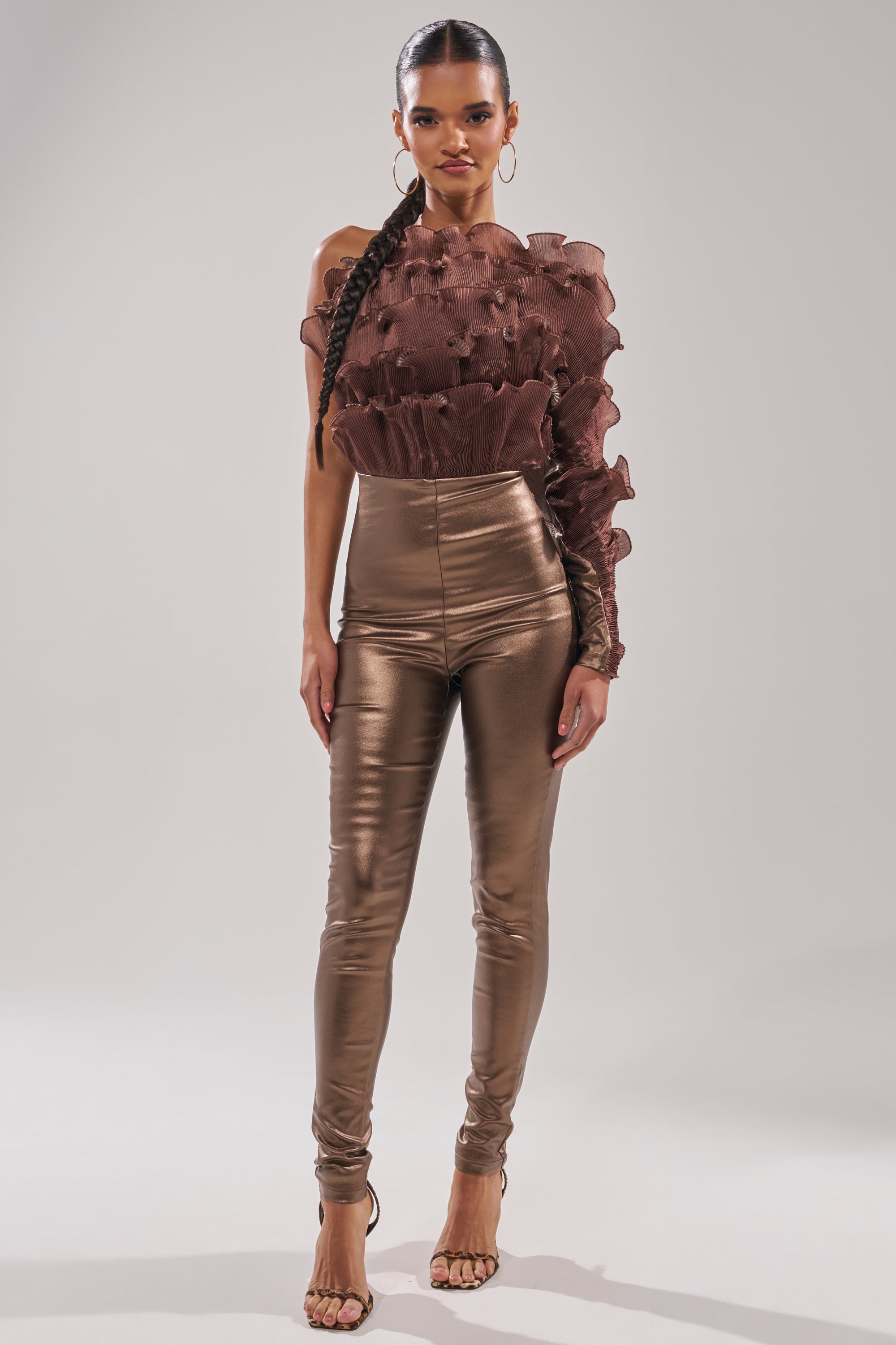 A woman faces forward in the COCOA BUTTER ONE SLEEVE JUMPSUIT, a textured brown metallic piece with one sleeve, paired with heels. Her hair is styled in a long, sleek braid against a plain light background.