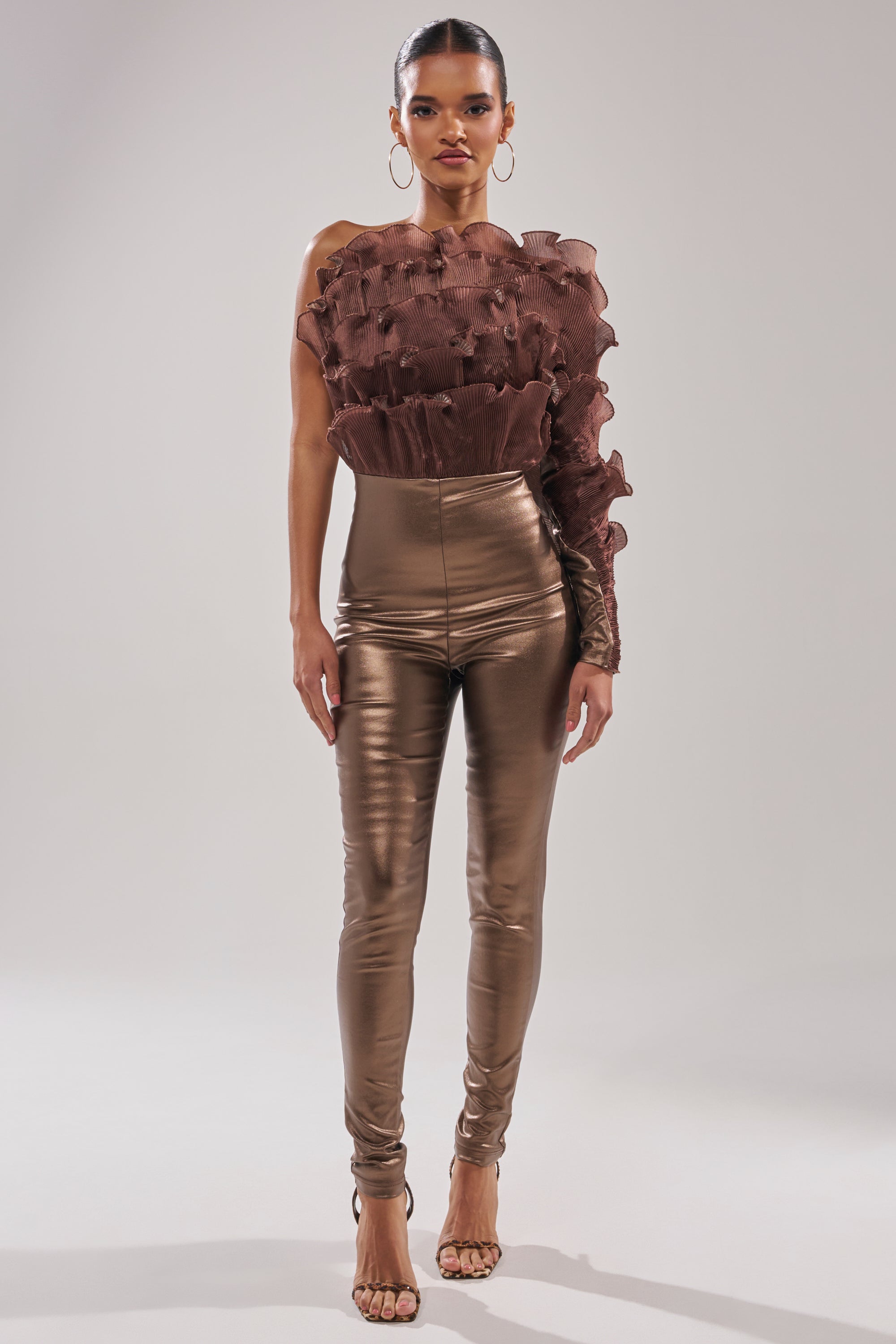 A woman wears the COCOA BUTTER ONE SLEEVE JUMPSUIT, featuring a textured, ruffled design, paired with shiny bronze high-waisted pants and open-toed heels against a plain light background.
