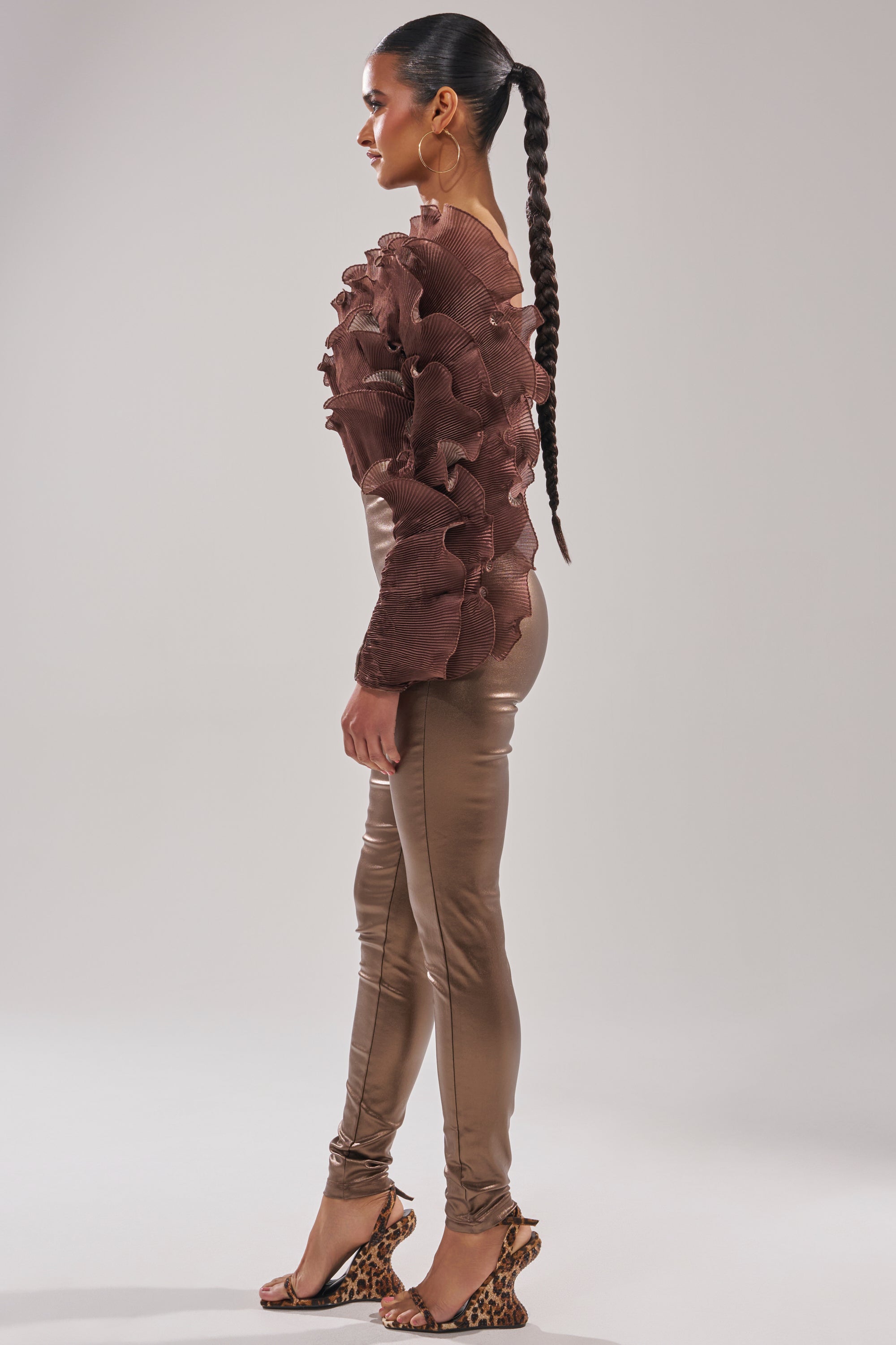 A woman stands in profile wearing the COCOA BUTTER ONE SLEEVE JUMPSUIT with a ruffled texture and shiny, fitted pants. She pairs it with a long braided ponytail and high-heeled leopard print sandals against a plain, light background.