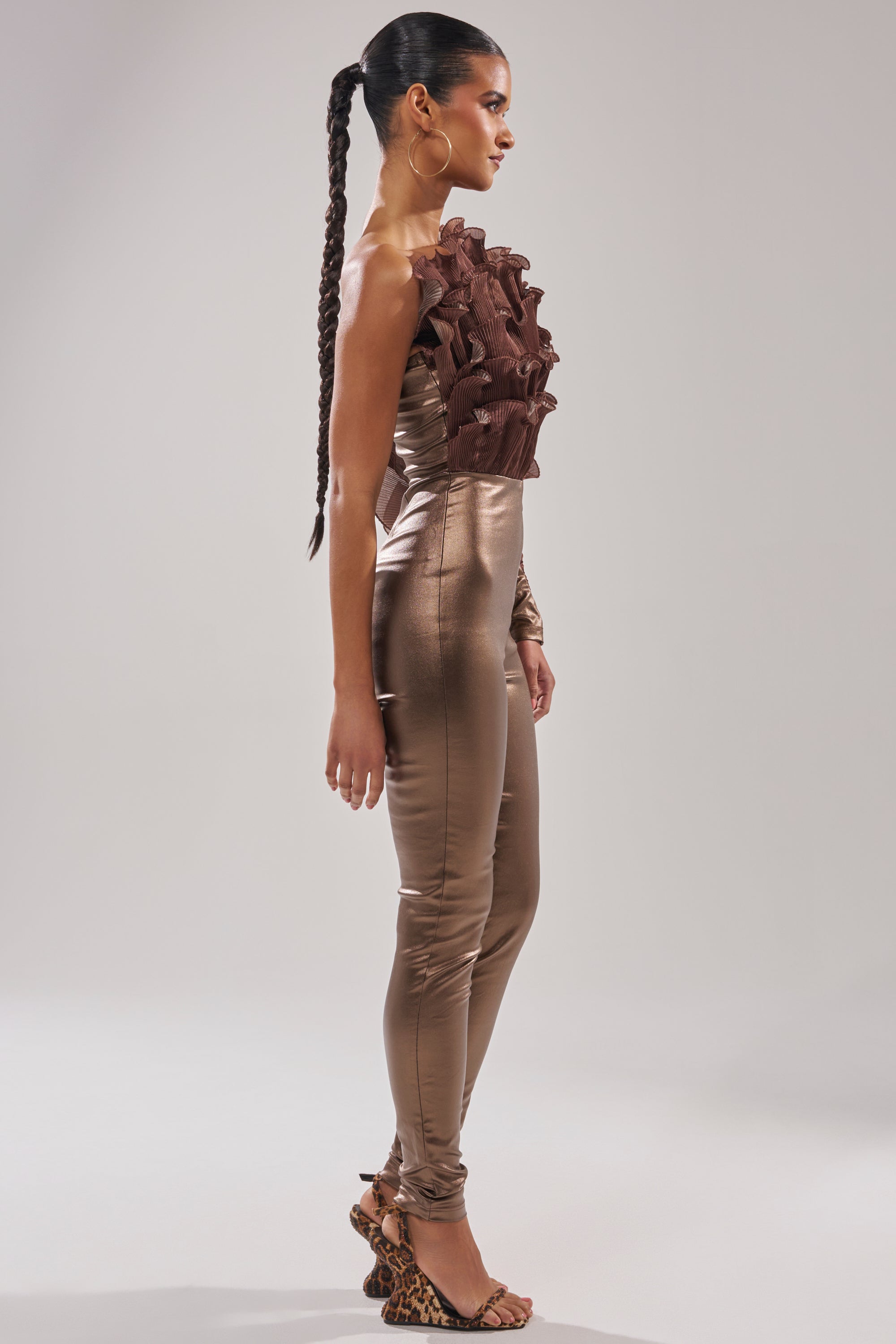 A woman poses in profile wearing the COCOA BUTTER ONE SLEEVE JUMPSUIT, featuring a sculptural textured brown ruffle top. She pairs it with a long braided ponytail, large hoop earrings, and leopard-print heeled sandals against a plain backdrop.