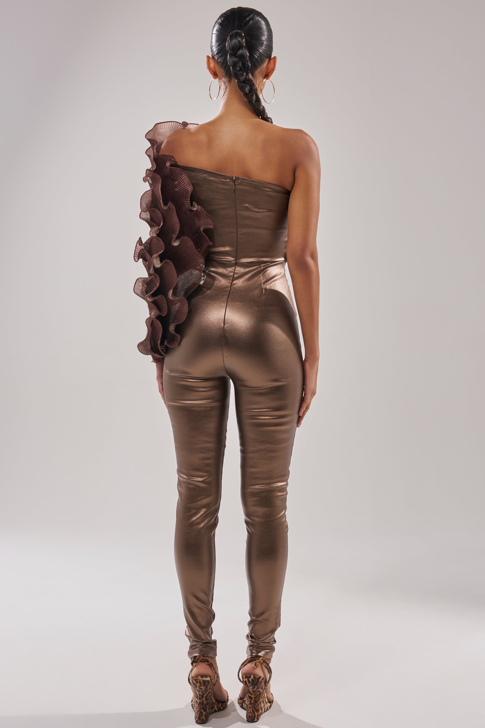 A woman stands facing away in the COCOA BUTTER ONE SLEEVE JUMPSUIT, a metallic bronze piece featuring a large ruffle on the left sleeve. Her hair is styled in a long braid, paired with strappy heels against a neutral background.