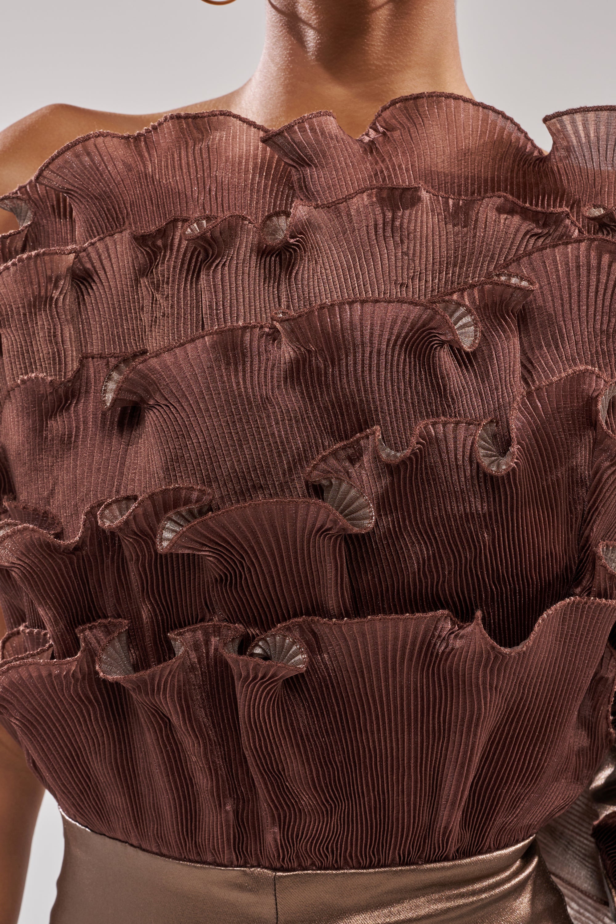 Close-up of a person wearing the COCOA BUTTER ONE SLEEVE JUMPSUIT, featuring dramatic, structured brown pleats and ruffled layers for a sculptural, artistic look. The textured fabric enhances its bold, statement style.