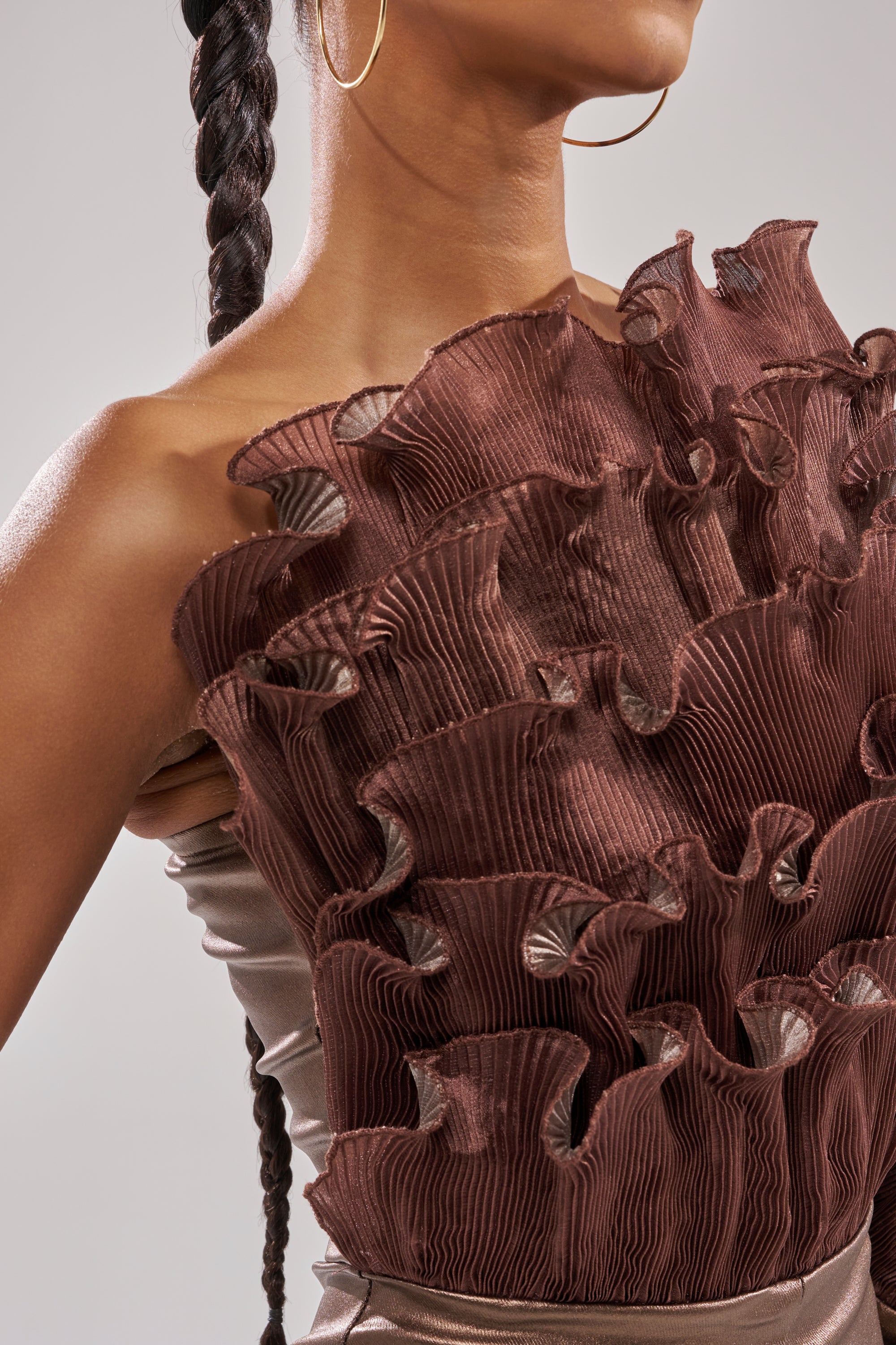 A woman with a long braid and hoop earrings wears the COCOA BUTTER ONE SLEEVE JUMPSUIT, designed with striking, textured brown ruffles across the chest and shoulder. The background is neutral.