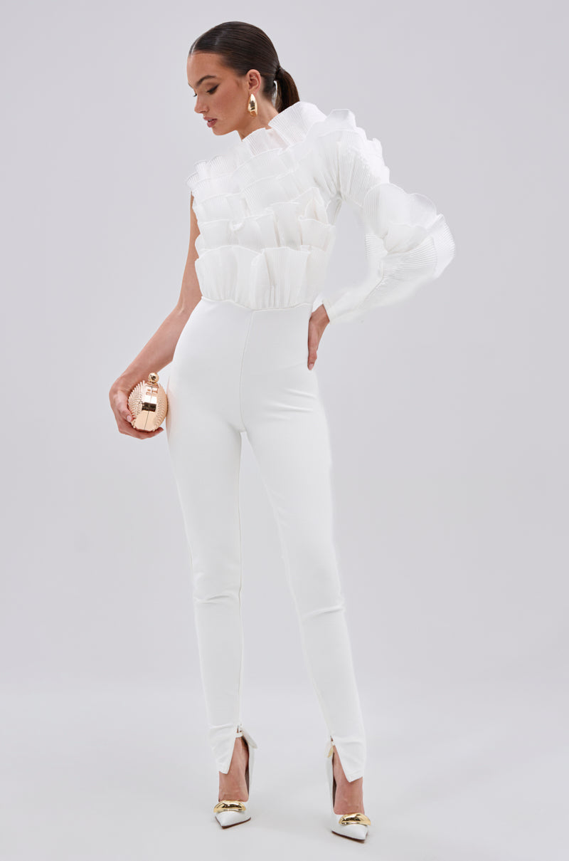 A woman models the ADA RUFFLE TOP BANDAGE JUMPSUIT with a bold mesh organza ruffle, paired with metallic gold heels and a round gold clutch, standing against a light gray background. 0
