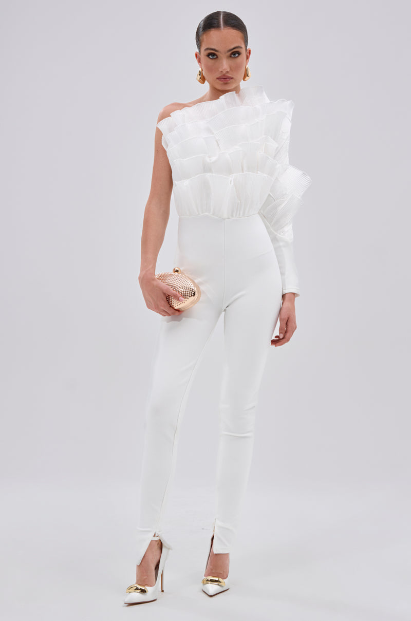 A woman stands against a plain background in the ADA RUFFLE TOP BANDAGE JUMPSUIT—an asymmetrical single-sleeve style with a mesh organza ruffle and fitted legs. She accessorizes with a metallic clutch and gold heels. 1