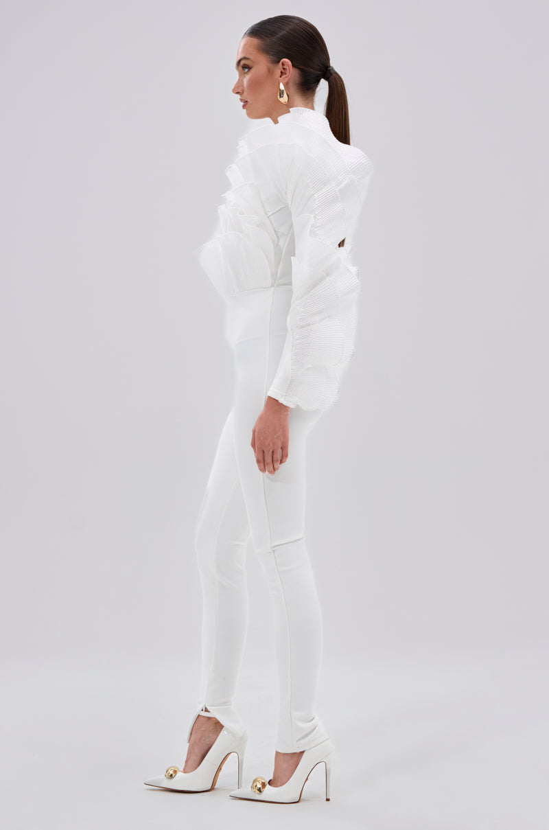 A woman in profile wears the ADA RUFFLE TOP BANDAGE JUMPSUIT featuring sculptural mesh organza ruffle sleeves and fitted pants, paired with white heels with gold accents. Her hair is slicked back and she accessorizes with large gold hoop earrings. 2
