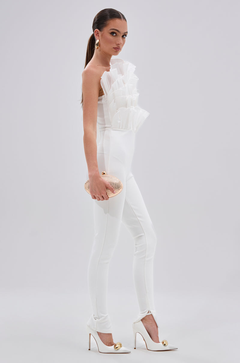 A woman in the ADA RUFFLE TOP BANDAGE JUMPSUIT, styled with sculptural mesh organza ruffles, fitted pants, white heels with gold accents, gold earrings, and a textured clutch bag stands against a plain light background. 3