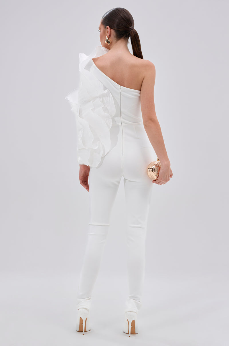 A woman stands facing away in the sleek white ADA RUFFLE TOP BANDAGE JUMPSUIT, holding a gold clutch. She has straight dark hair in a low ponytail, large gold earrings, and wears high heels. 4