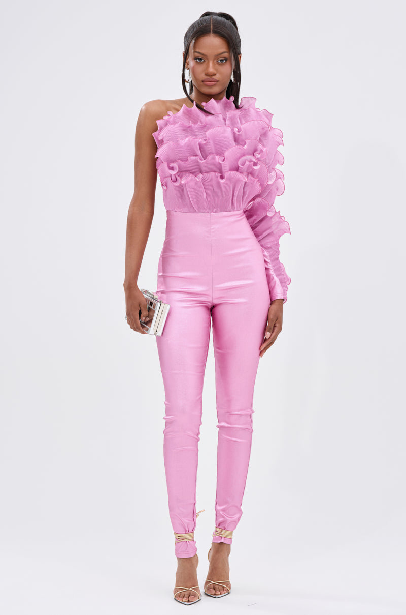 A woman stands against a plain background wearing the RAQUEL RUFFLED JUMPSUIT, a shiny magenta one-sleeve jumpsuit with ruffled detailing. She accessorizes with a small clutch, strappy heels, and a half-up, half-down hairstyle.