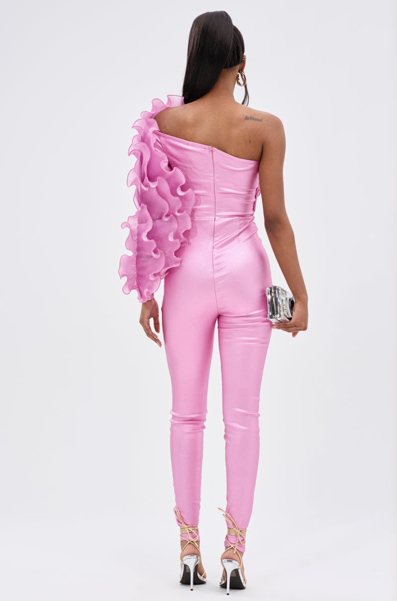 A woman faces away in the RAQUEL RUFFLED JUMPSUIT—a shiny magenta piece with a dramatic ruffled sleeve. She pairs it with strappy heels, a silver clutch, straight hair, and large hoop earrings.