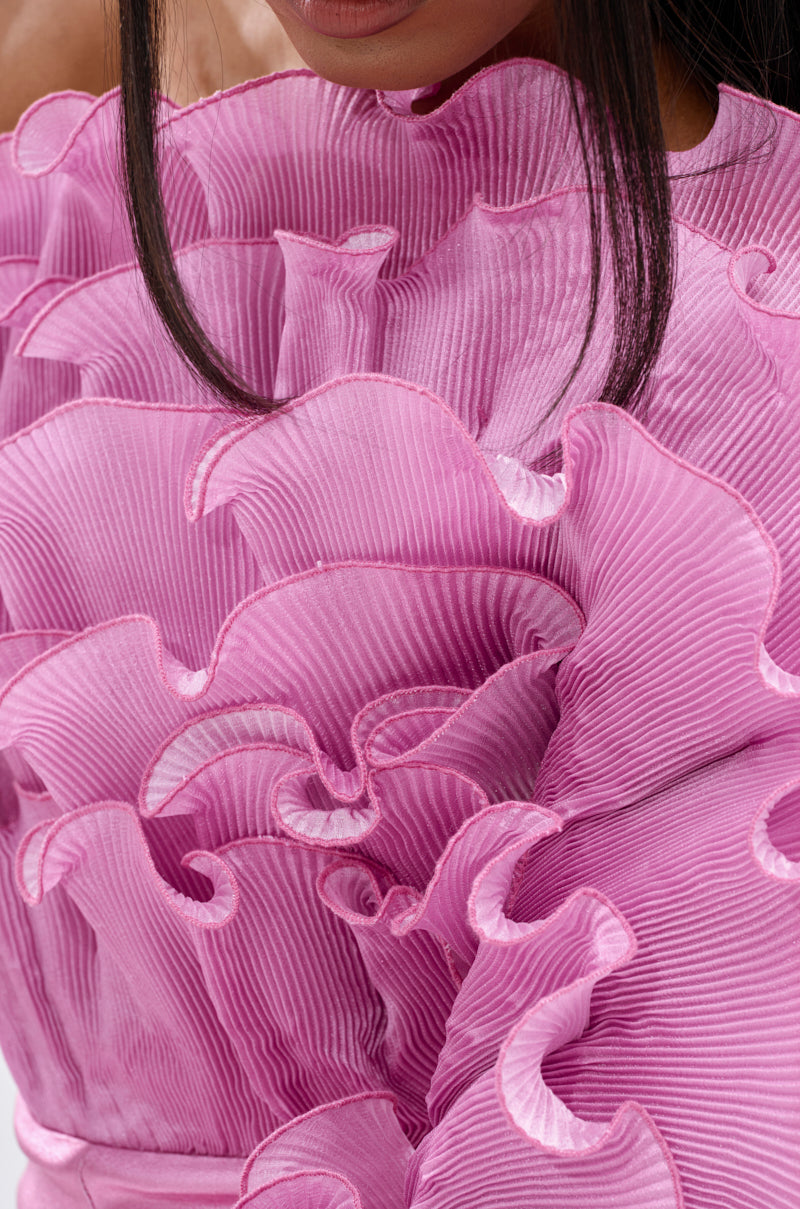Close-up of a person wearing the RAQUEL RUFFLED JUMPSUIT, featuring sheer pink fabric with wave patterns and sculptural, layered ruffles. Only the lower face and upper body are shown.