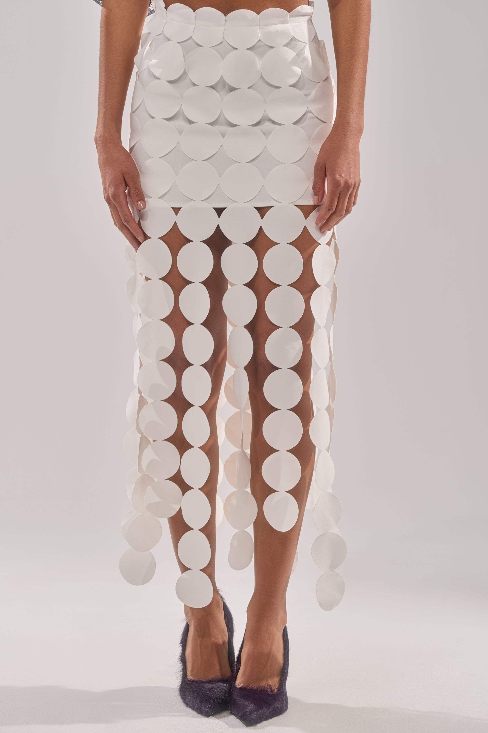 A person wears the ROYALTY CIRCLE CUT OUT MAXI SKIRT, featuring large white circular discs and fringe-like hanging circles, paired with dark pointed shoes. Only the lower half of the body is shown. 1