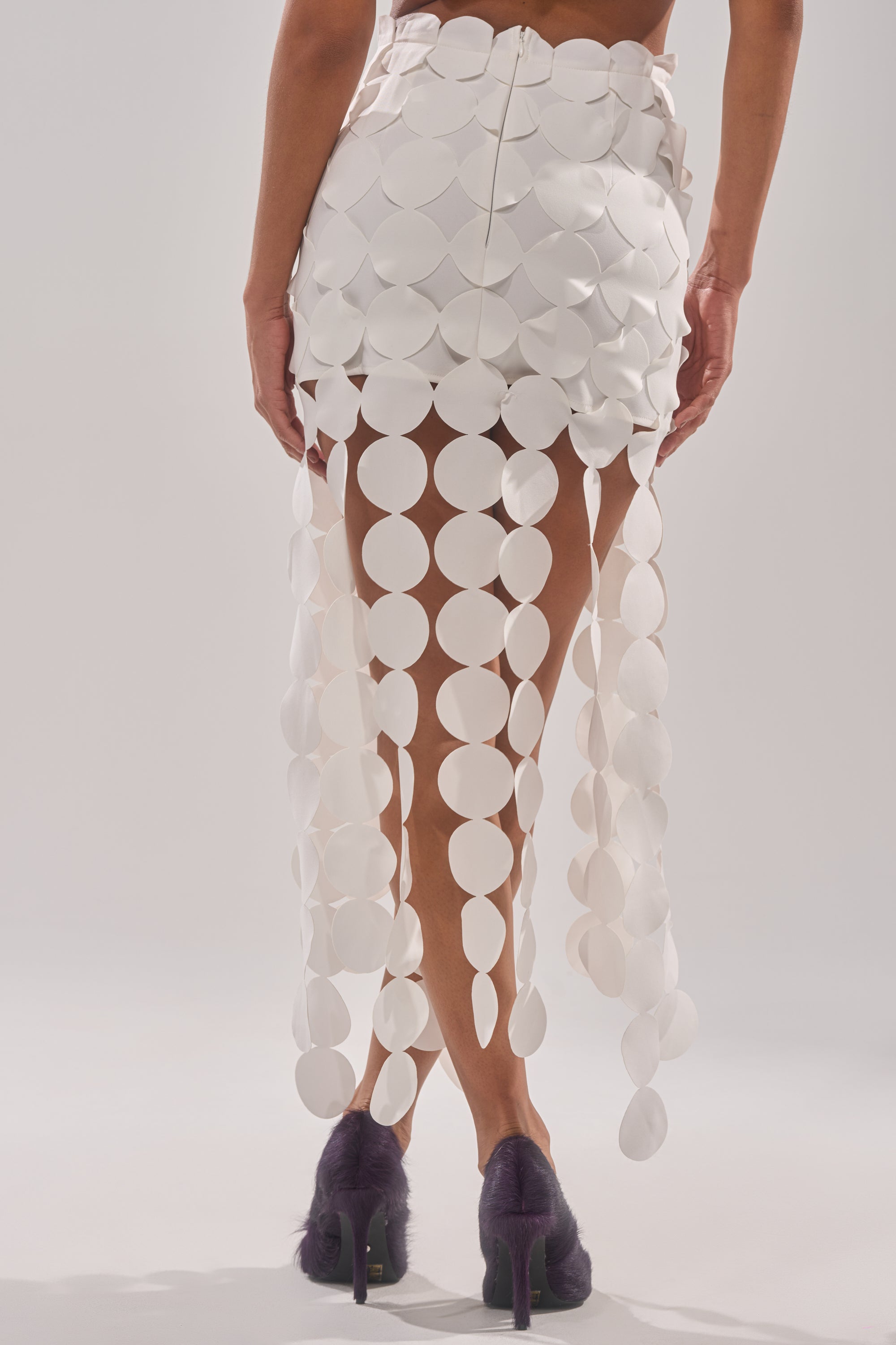 Wearing the ROYALTY CIRCLE CUT OUT MAXI SKIRT, featuring strands of large white discs, paired with furry dark high-heeled shoes, a person stands against a plain light background. 3