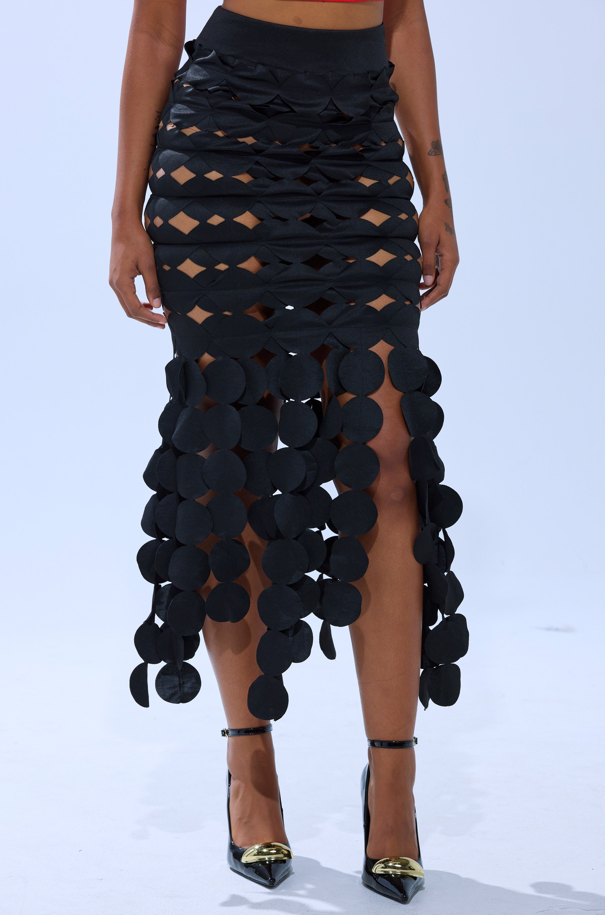 A person wears the LOOKING FOR ME CIRCLE CUTOUT SKIRT with diamond-shaped cutouts and circular accents at the hem, paired with black stiletto heels featuring metallic gold pointed toes. 1