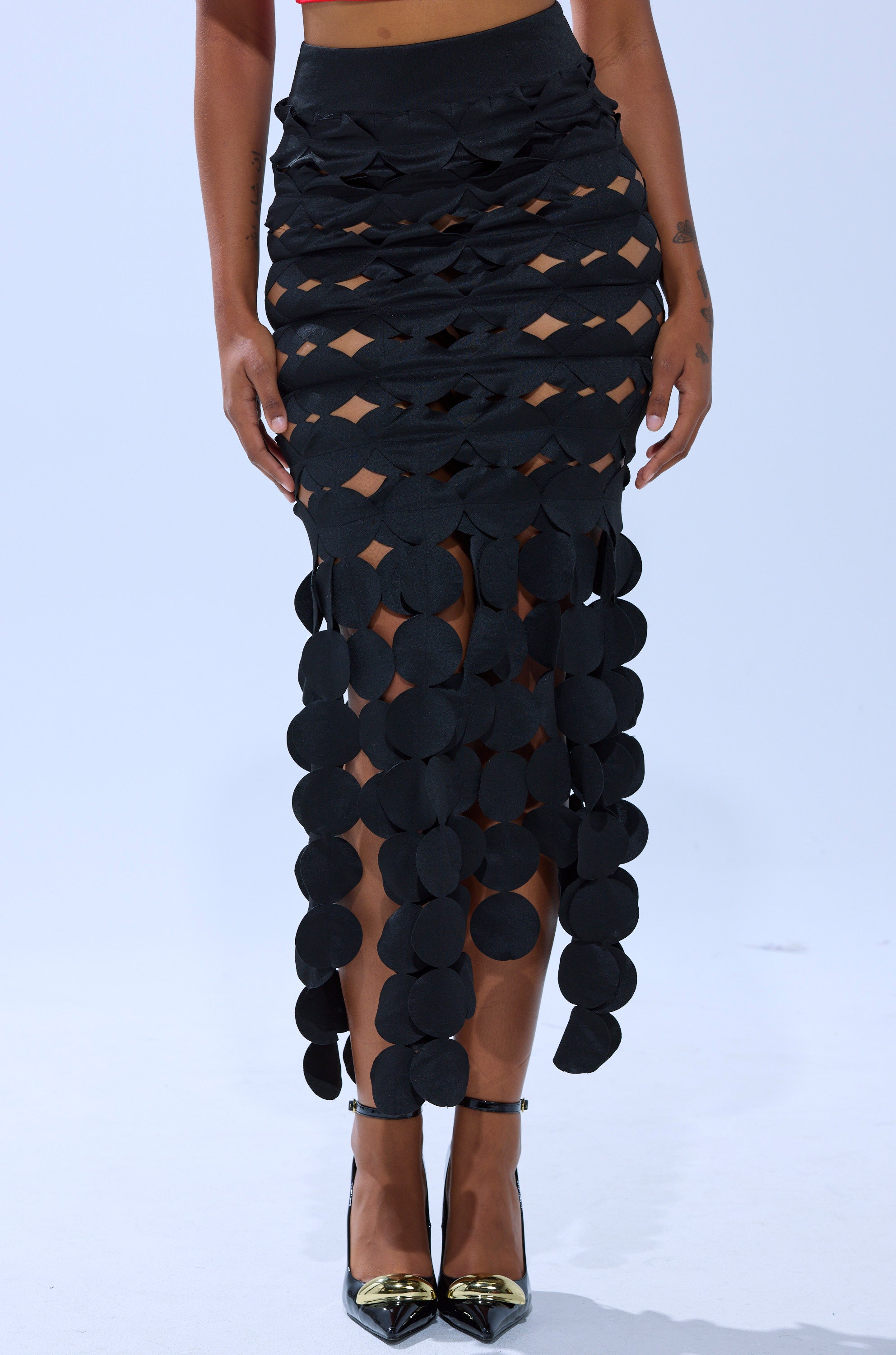 Someone wears the LOOKING FOR ME CIRCLE CUTOUT SKIRT, a distinctive black maxi with interlacing bands and cascading circles, styled with black high heels against a plain light background. 2