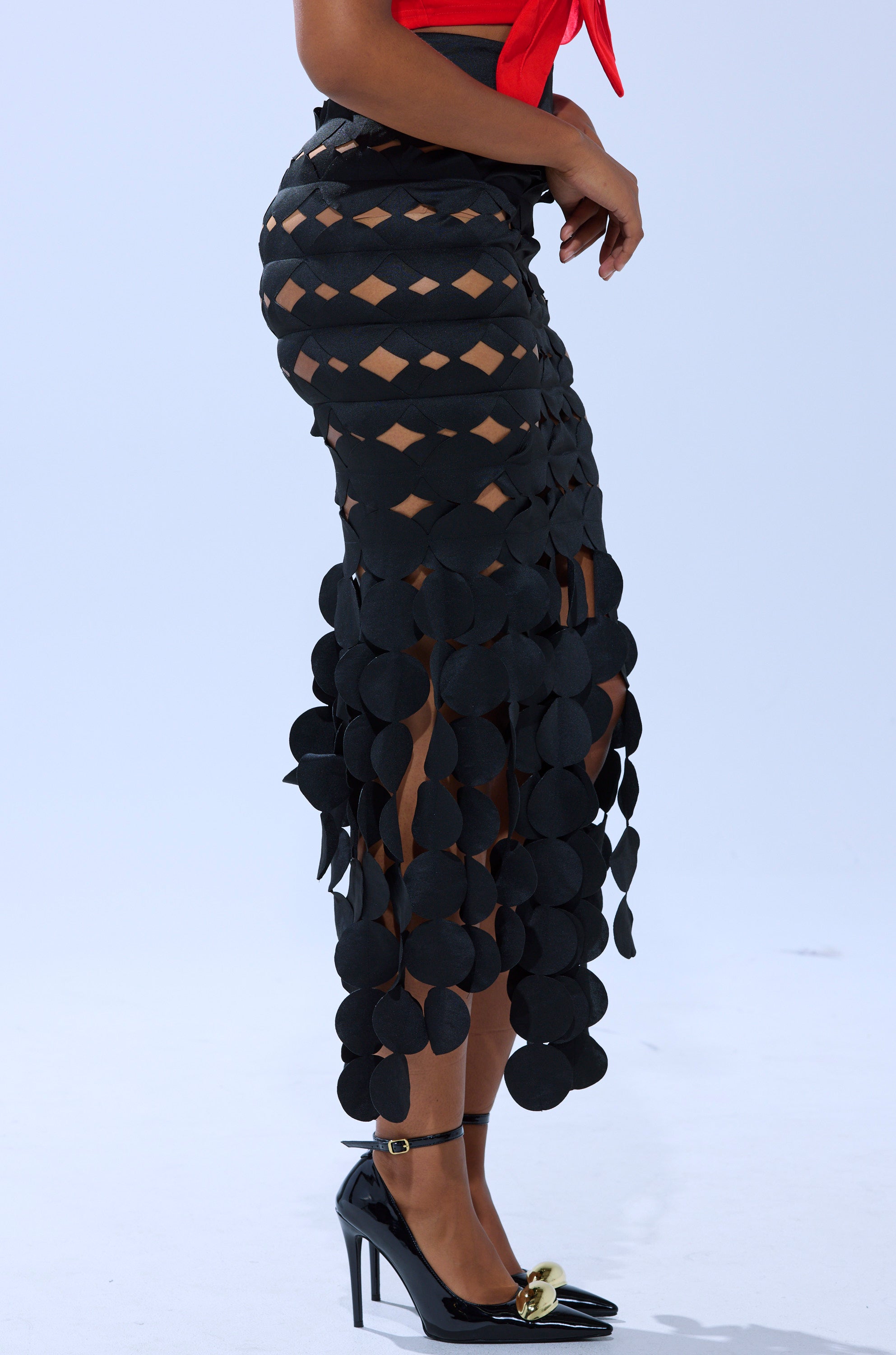 A person wears the LOOKING FOR ME CIRCLE CUTOUT SKIRT in black, featuring skin-revealing cutouts, paired with black ankle-strap high heels and a partially visible red top. 3