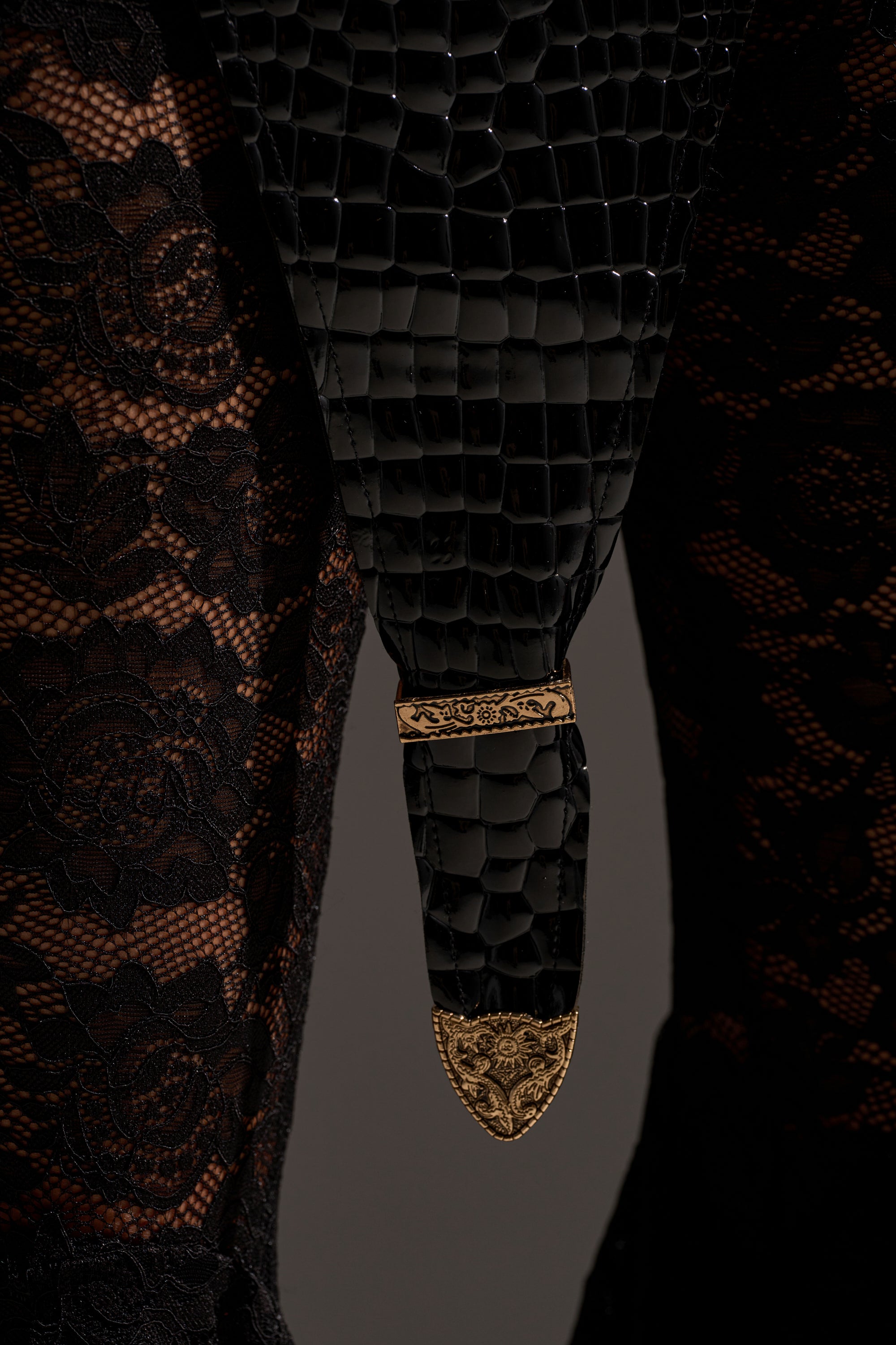 Close-up of a person wearing the BUCKLE DOWN CORSET, featuring black floral lace fabric, faux leather with crocodile embossing, and ornate gold buckle details on the tip and buckle.