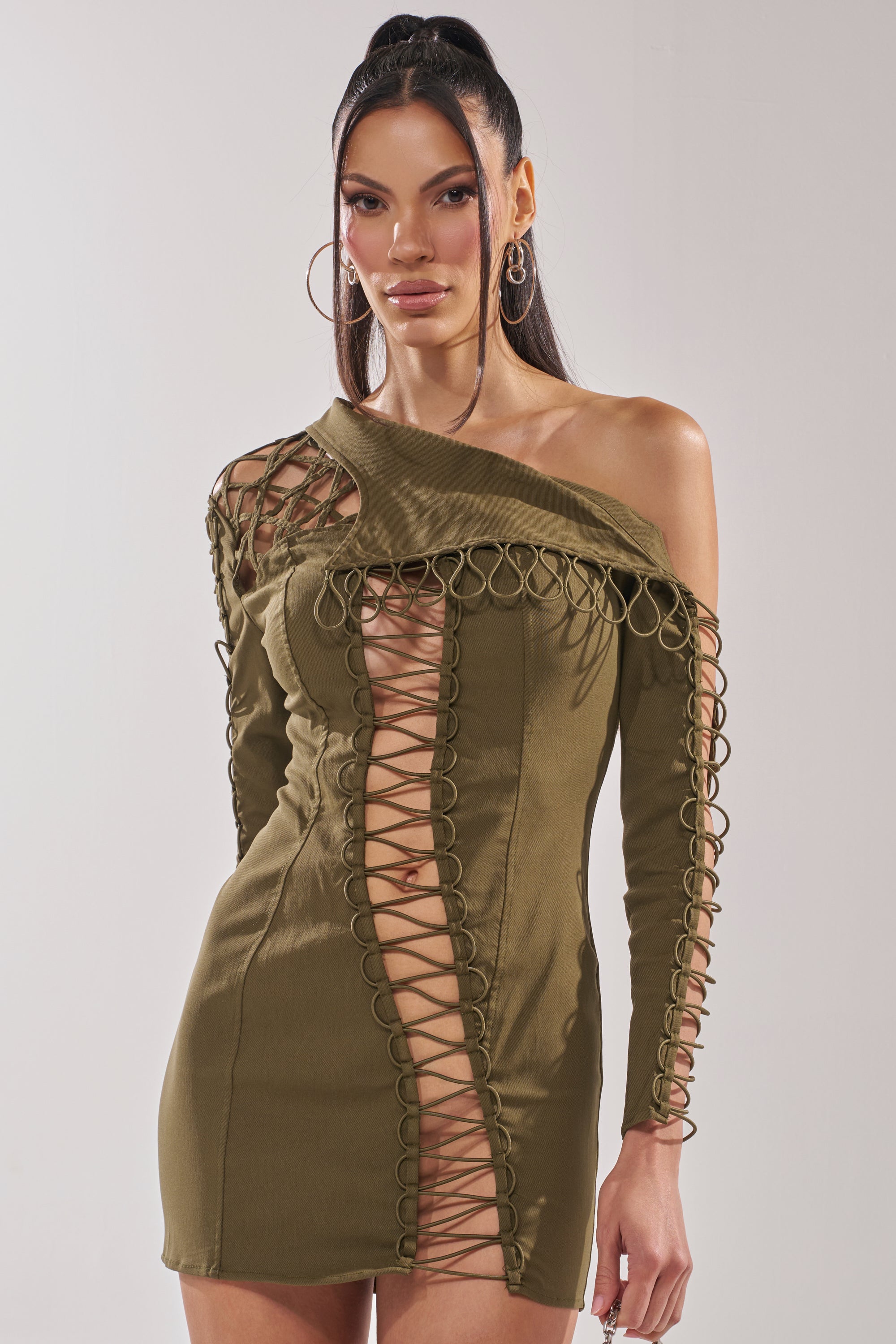 A woman models the LACED UP MINI DRESS—an olive one-shoulder mini with looped trim and a striking lace-up cutout running diagonally from shoulder to hem—against a plain light background. 2