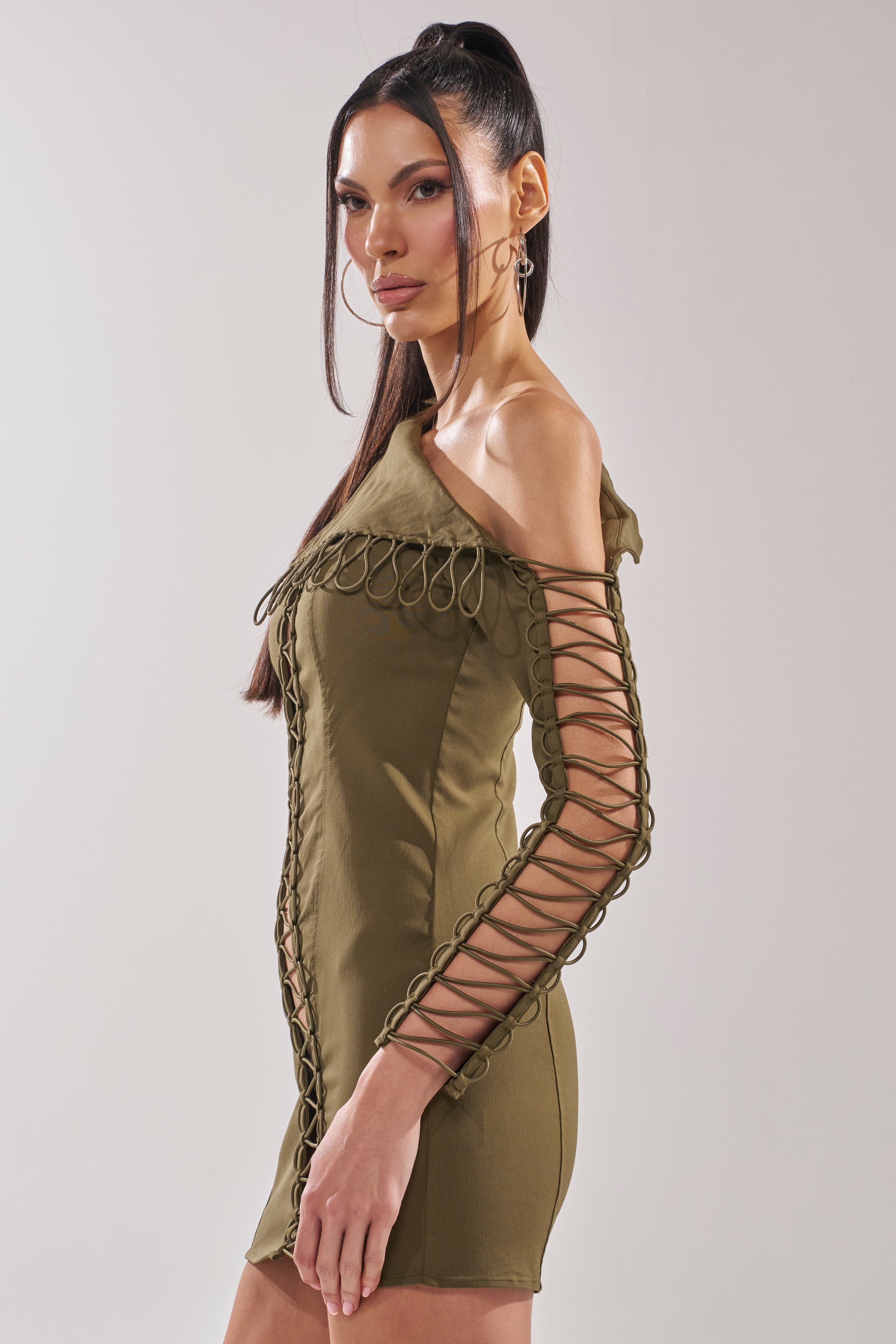 Woman wearing the LACED UP MINI DRESS in olive, featuring a one-shoulder design, decorative loops, and lace-up cutouts along the sleeves and front, posed against a light neutral background. 4