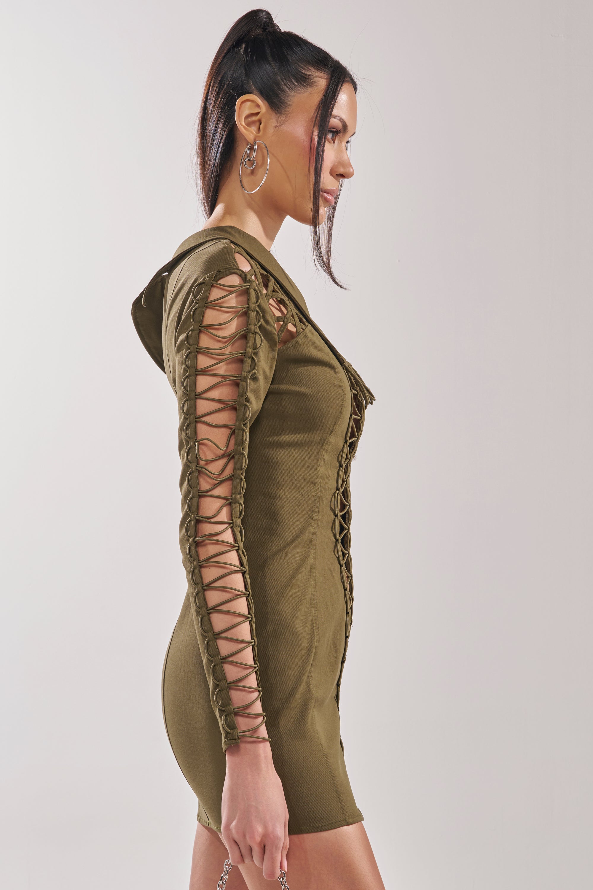 A woman in profile wears the LACED UP MINI DRESS, a fitted bodycon club dress with long sleeves and crisscross lace-up arm details. She pairs it with hoop earrings and a high ponytail for a chic, confident look. 5