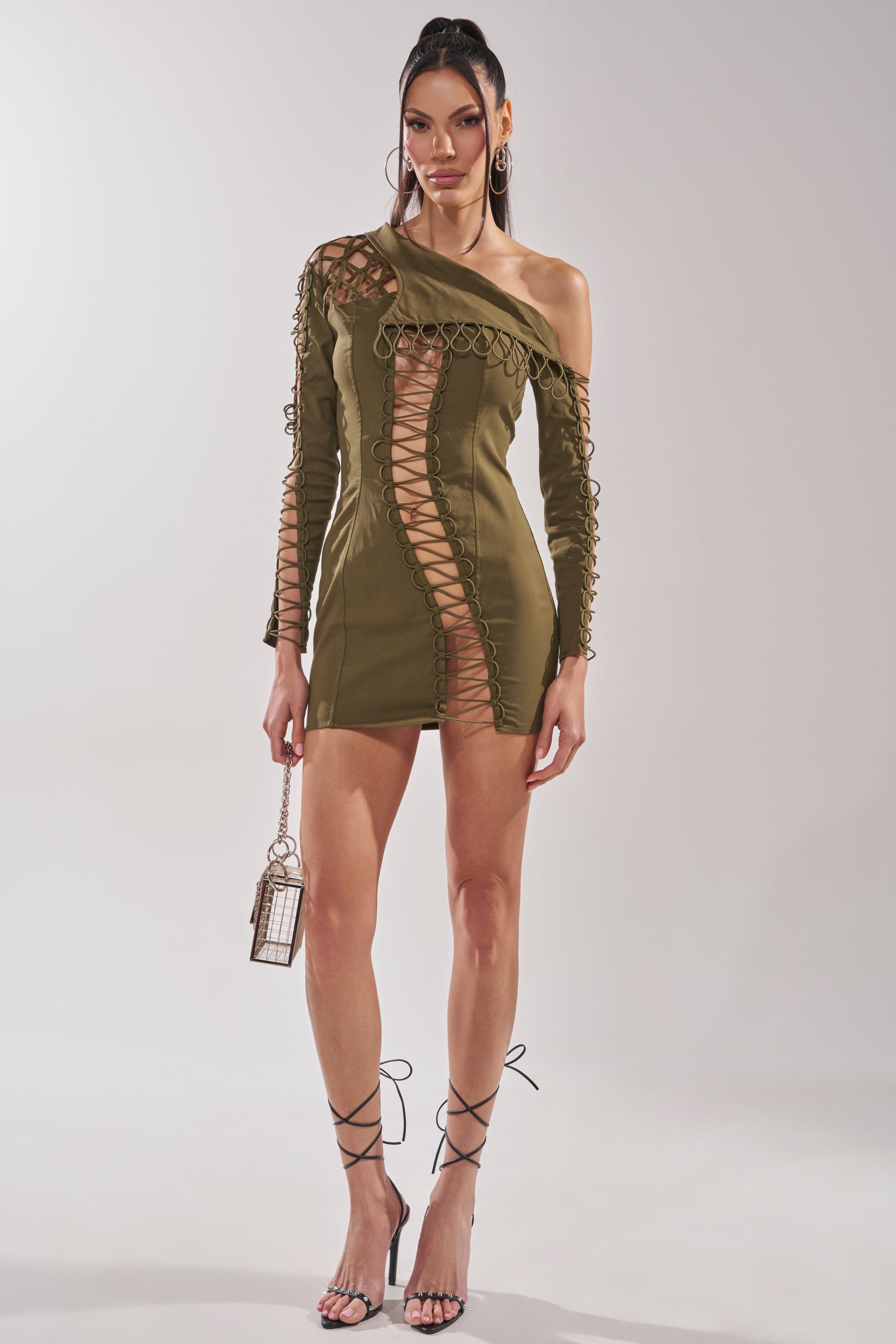 A woman wears the LACED UP MINI DRESS, an olive one-shoulder mini with lace-up design and a single long sleeve, styled with a small bag and strappy high heels against a plain background for a bold party look. 7