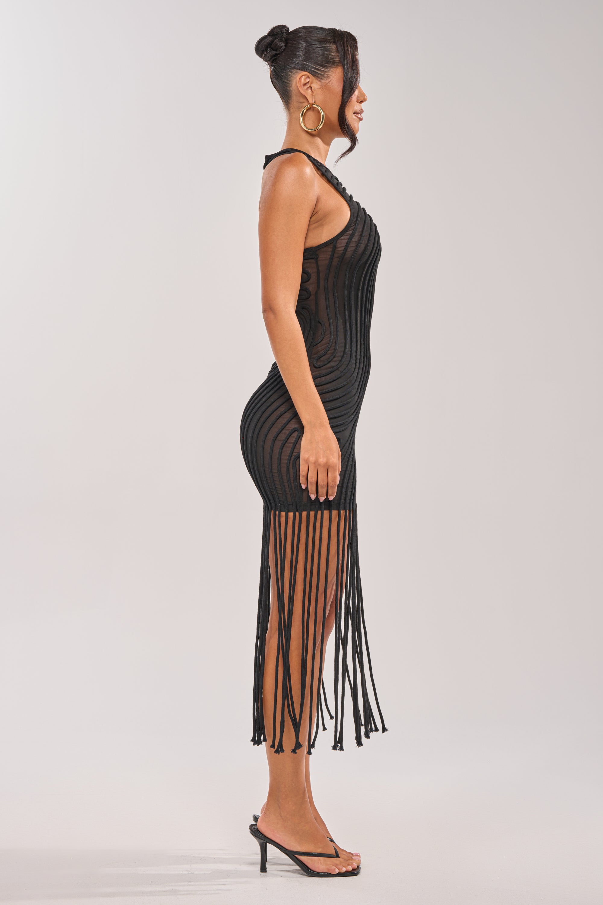 A woman stands in profile wearing the ILLUSIONS SLEEVELESS MINI DRESS IN BLACK, featuring vertical sheer mesh stripes and a fringe hem, styled with black strappy heels, gold hoop earrings, and a high bun. 4