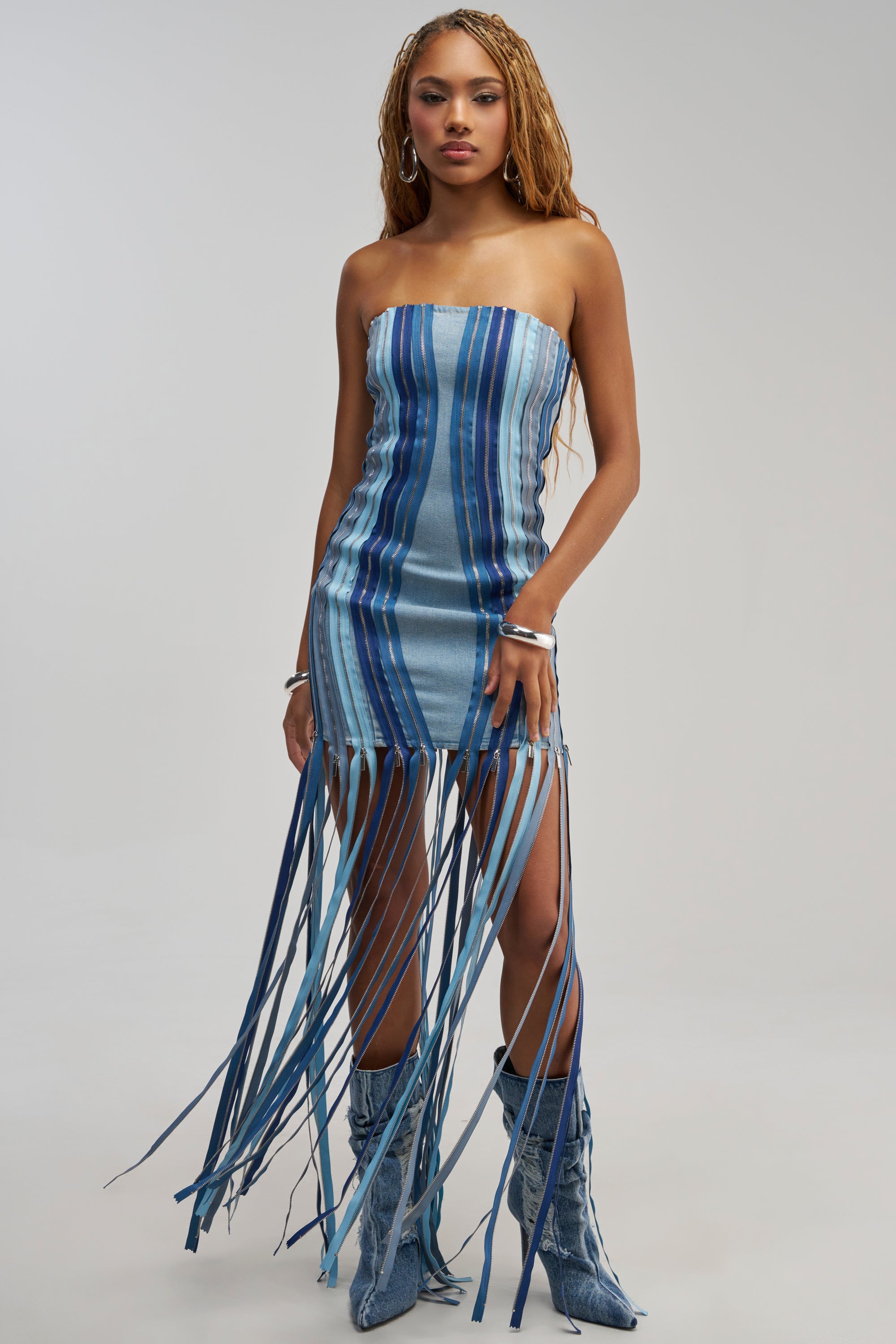 Wearing the DID IT AGAIN DENIM MINI DRESS IN MULTI—a strapless, blue striped denim mini dress with long fringe at the hem—paired with matching blue boots, a woman stands against a plain background, effortlessly channeling Y2K streetwear vibes.