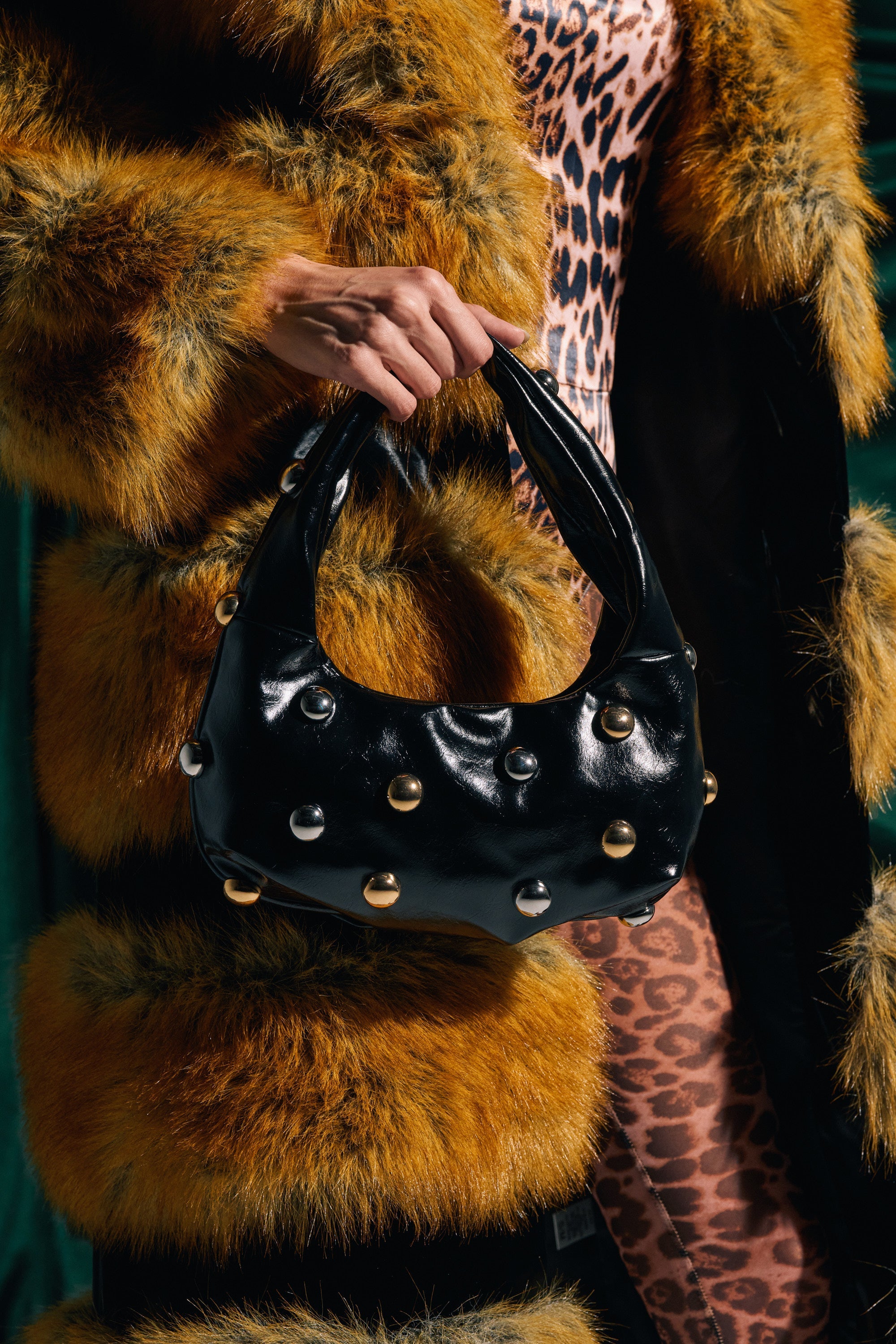 A person in a leopard print outfit and faux fur coat holds the TOO MOD BALL PURSE, featuring a shiny black surface decorated with large silver studs. 0